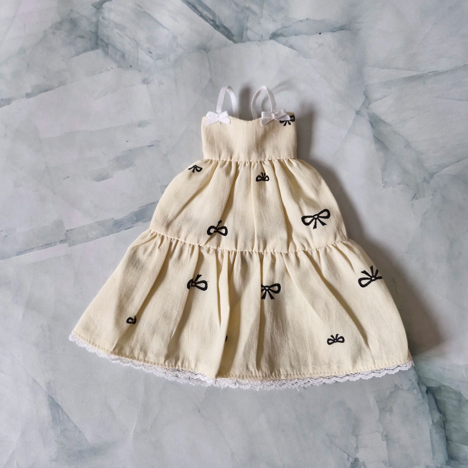 Cream tiered doll sundress with black bow print, white lace hem, and strap bows, laid flat on marble background.