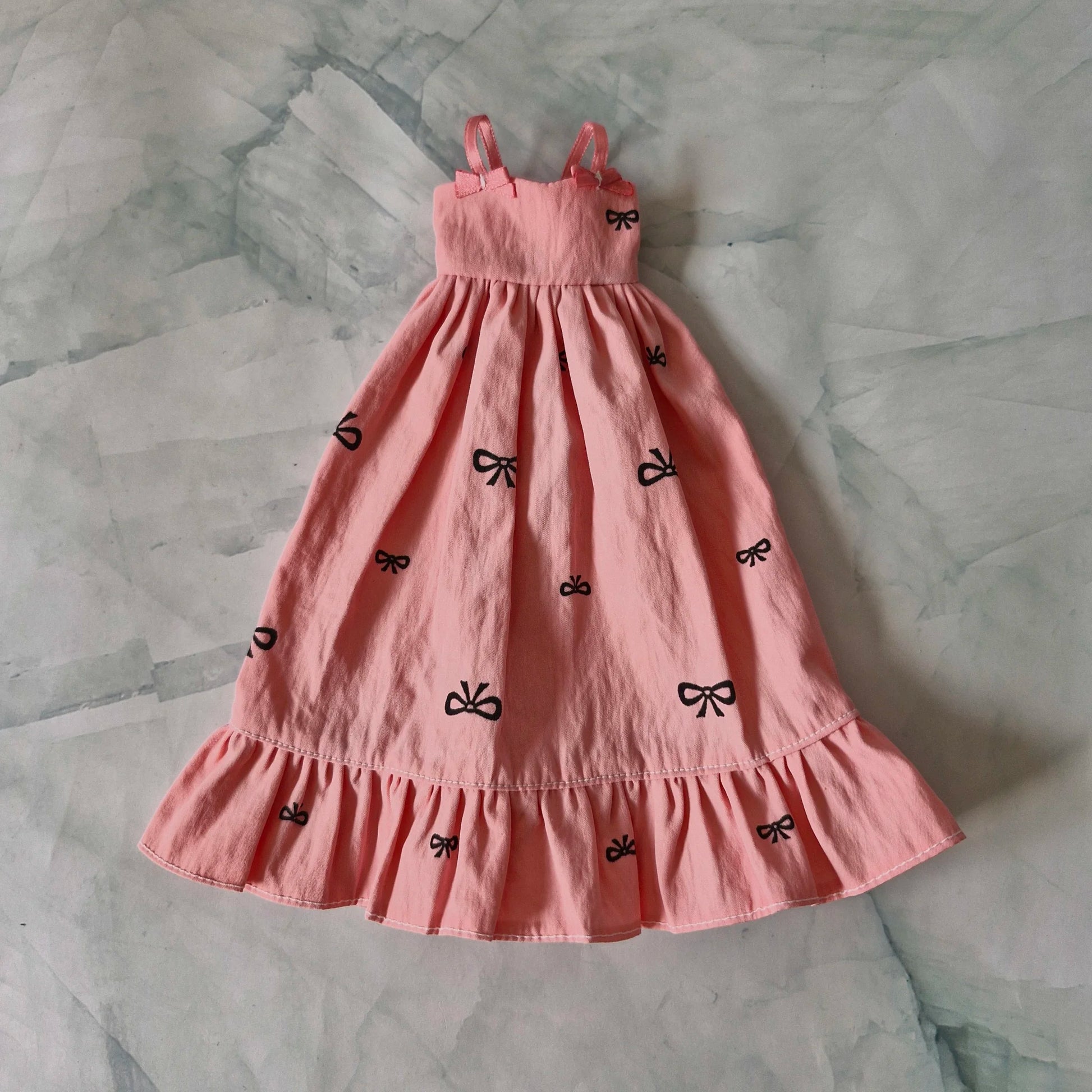 Pink doll sundress with black bow print, ruffle hem, and strap bows, laid flat on marble background.