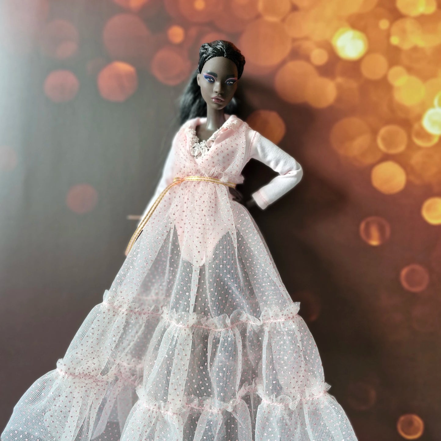 Close-up of dark-skinned Barbie doll in pink tiered sparkle tulle gown with gold belt, dark bokeh background.