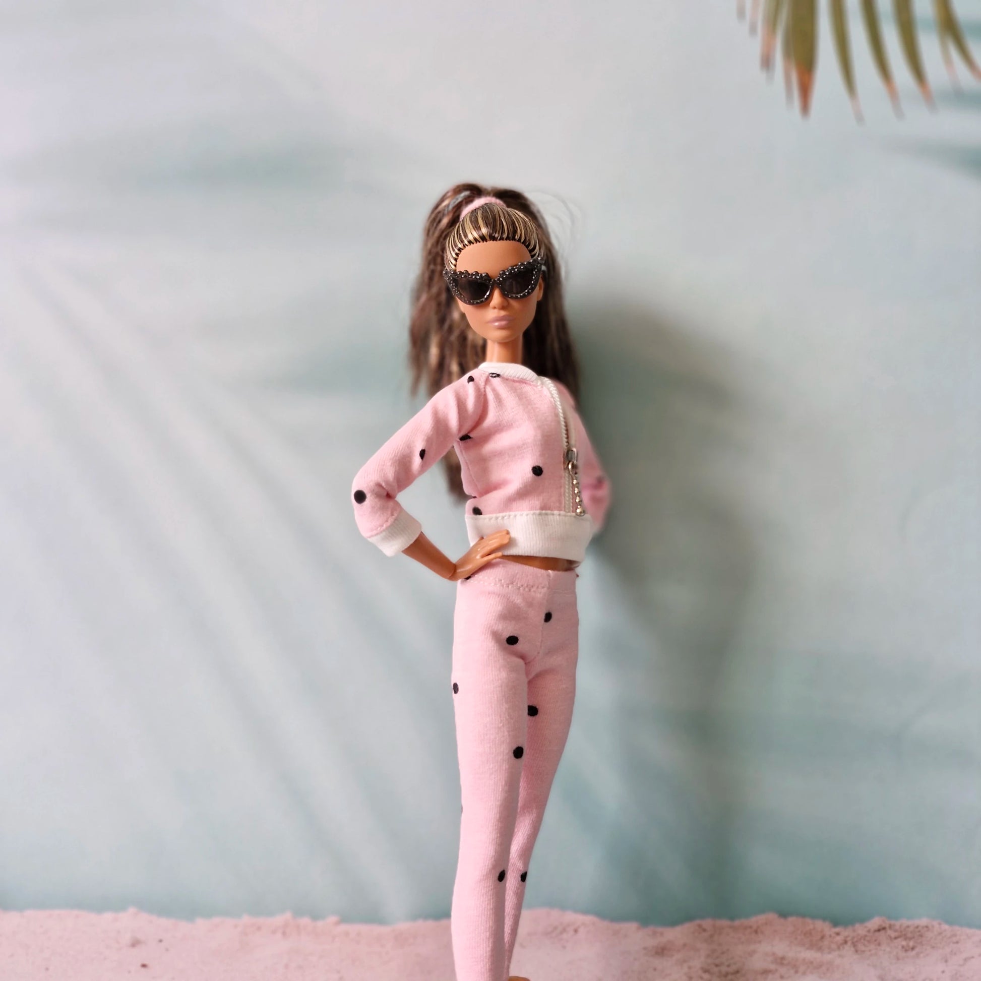 Barbie doll wearing pink polka dot tracksuit and stylish sunglasses, beach backdrop.