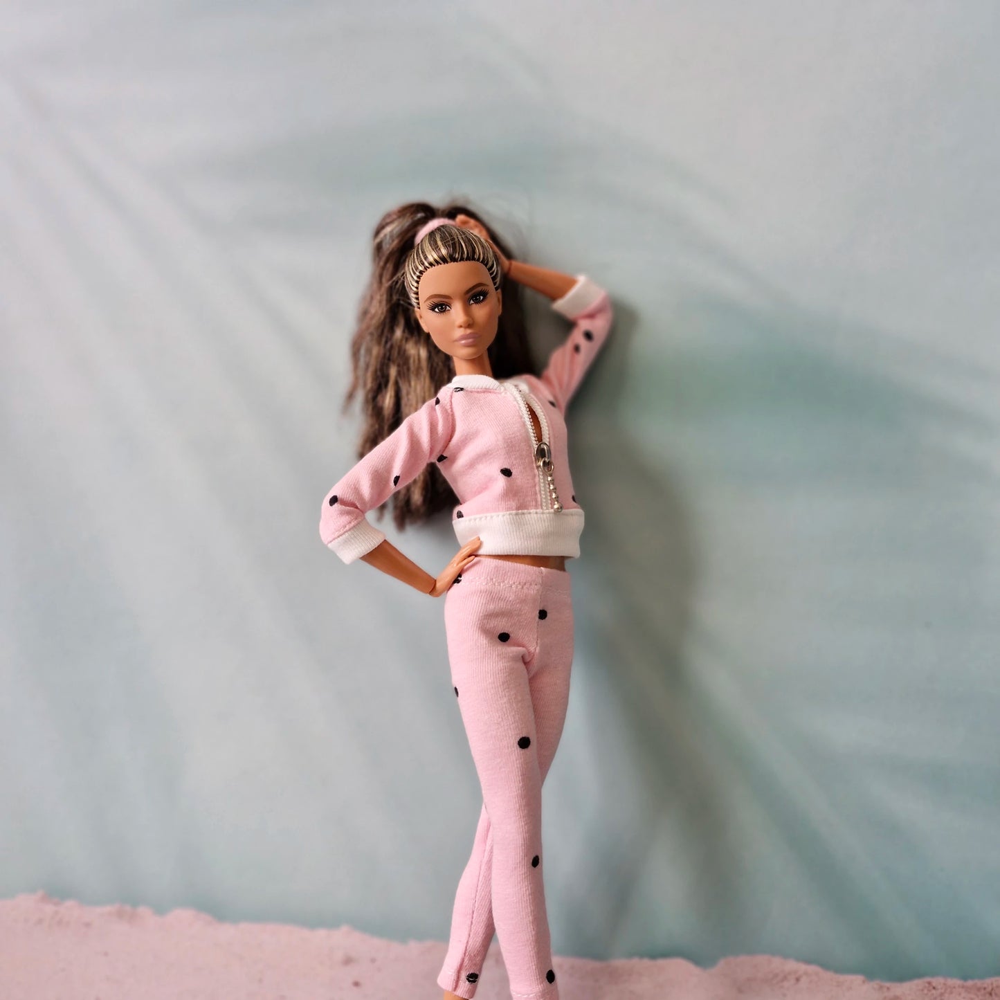 Barbie doll models pink polka dot tracksuit, hand behind head, beach theme.