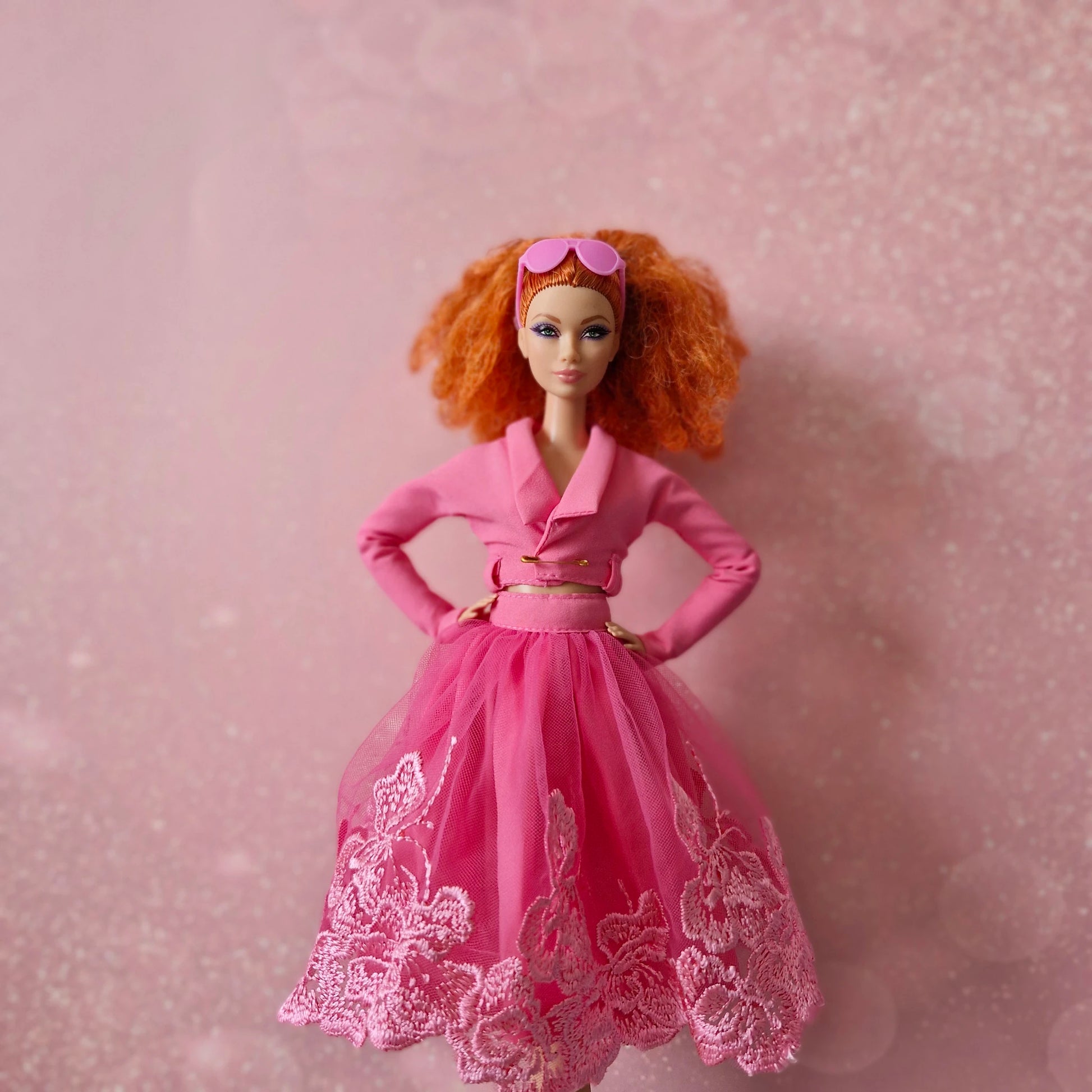Red-haired Barbie doll in pink cropped jacket and pink lace tulle skirt, sunglasses on head.
