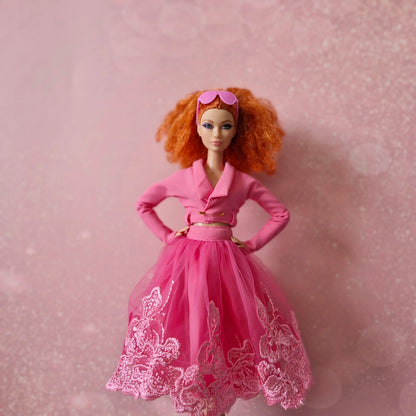 Red-haired Barbie doll in pink cropped jacket and pink lace tulle skirt, sunglasses on head.
