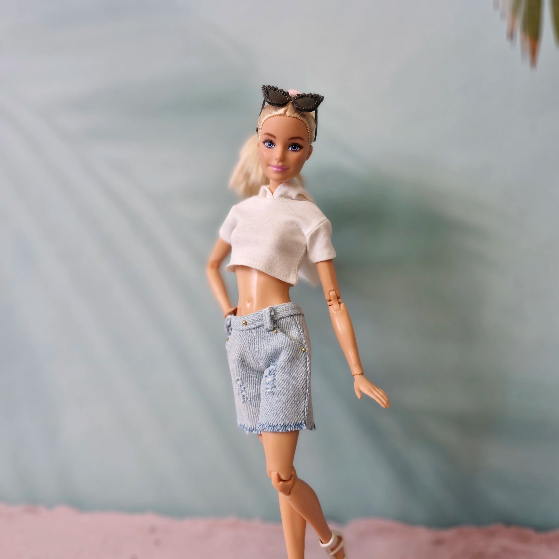Barbie doll with blonde hair in white crop top and light wash denim shorts, cat-eye sunglasses.