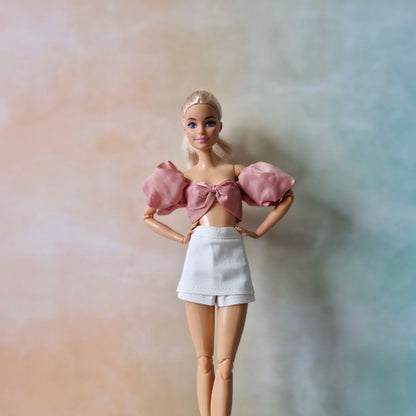 Barbie doll with blonde hair wearing stylish dusty rose puff sleeve bow front top and white mini skirt, hands on hips.