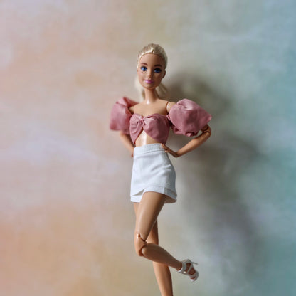 Blonde Barbie doll posing in dusty rose puff sleeve bow crop top and white mini skirt with white heels.