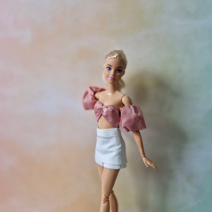 Blonde Barbie doll models dusty rose off-shoulder puff sleeve top with bow and white mini skirt.