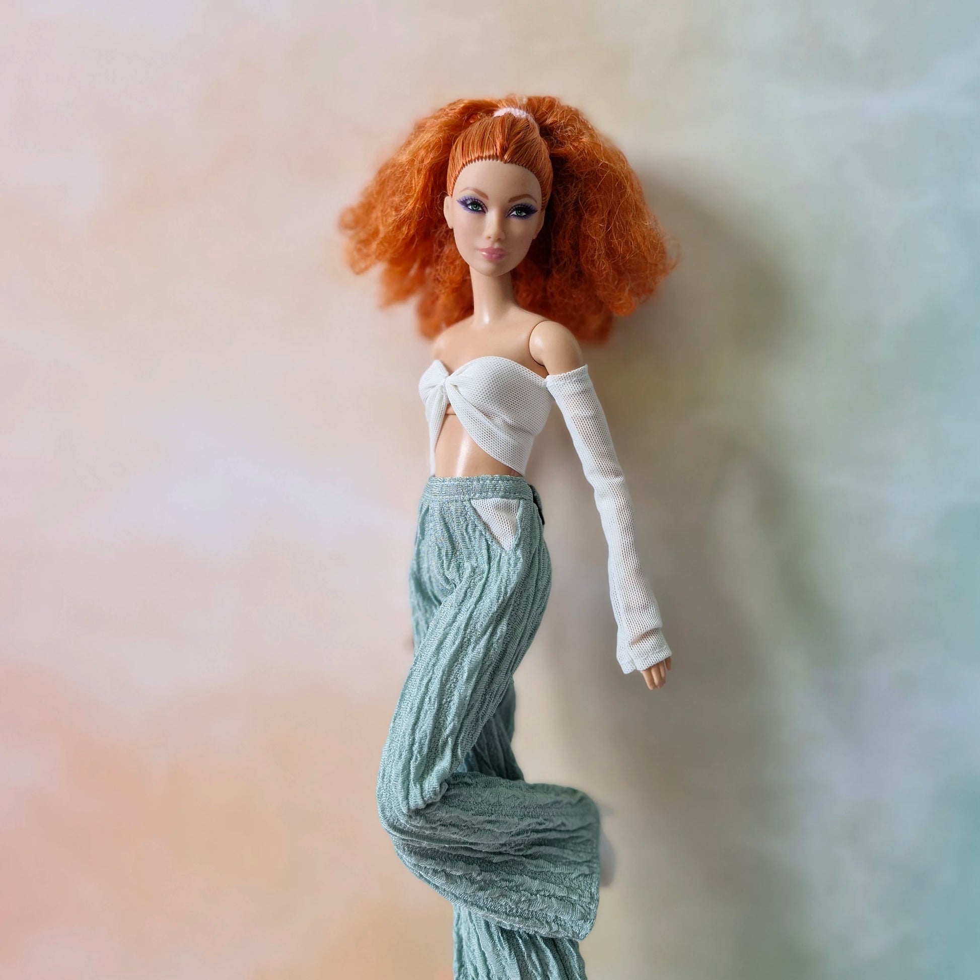Red-haired Barbie doll striding in white mesh sleeve crop top and flowy mint green textured pants.