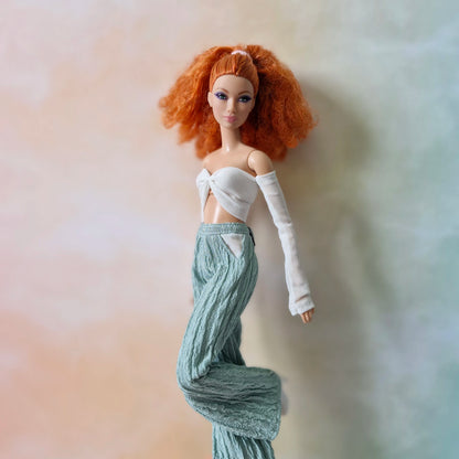 Red-haired Barbie doll striding in white mesh sleeve crop top and flowy mint green textured pants.