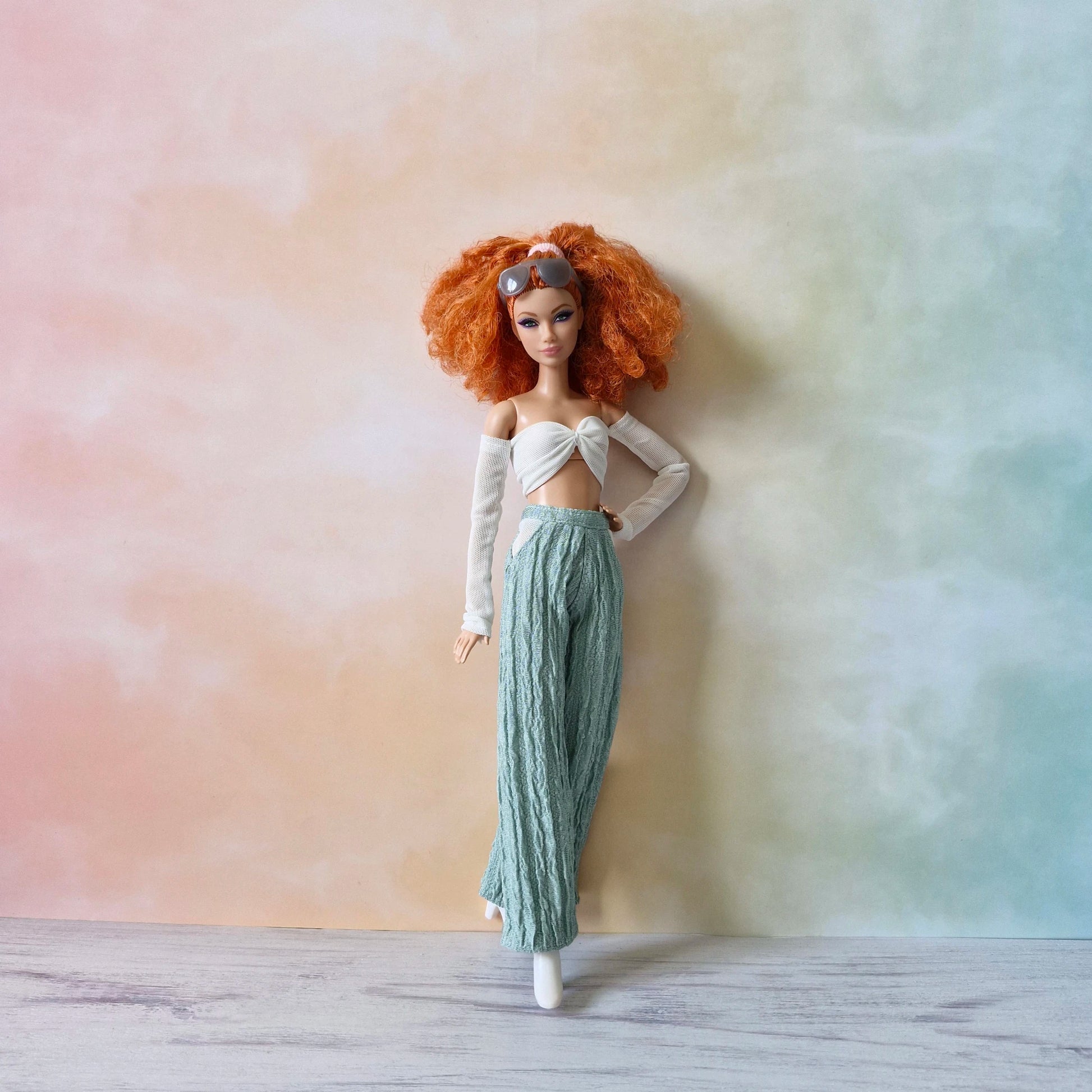 Barbie doll with sunglasses on head, white twist top, and mint green textured wide-leg pants, leaning casually.