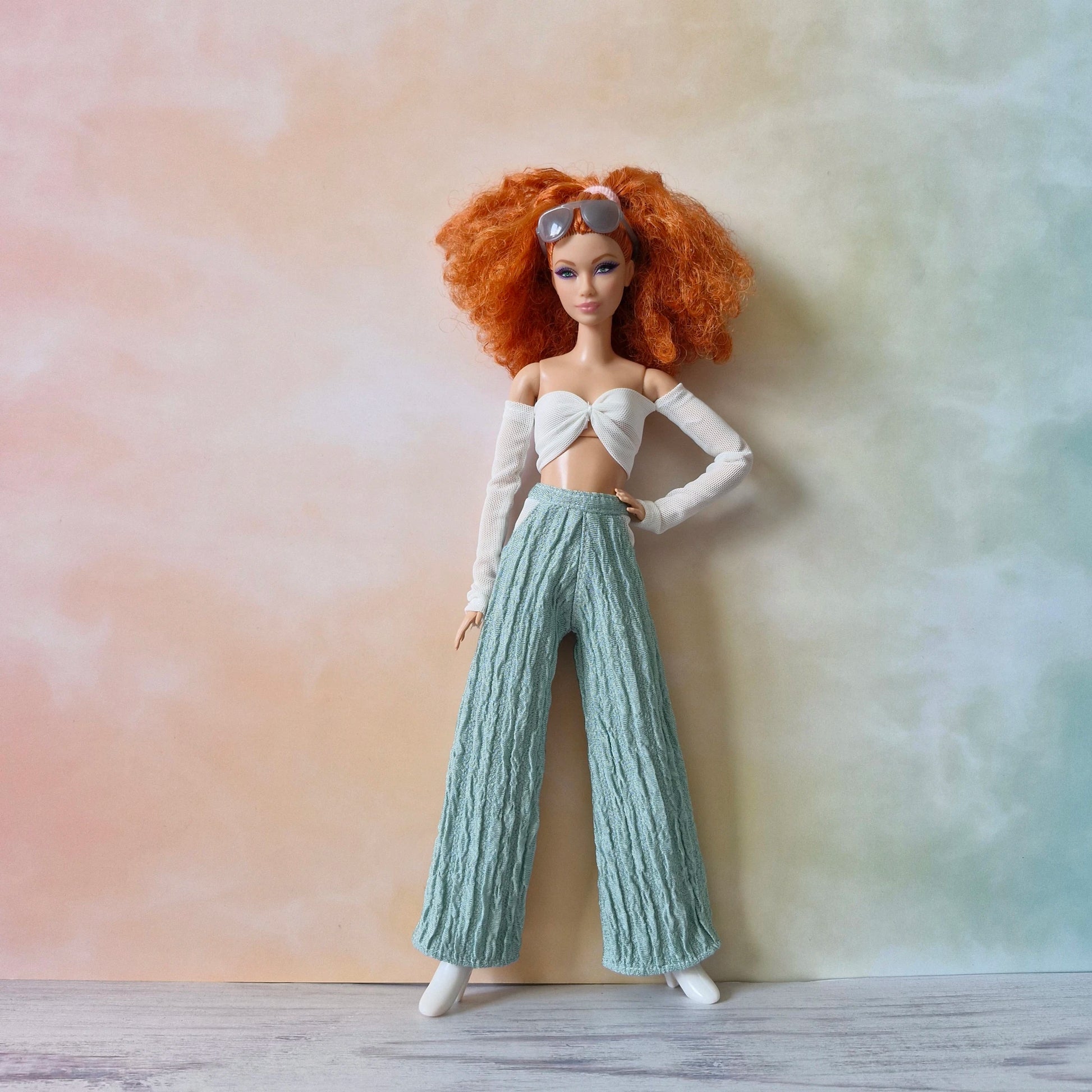 Red-haired Barbie doll with sunglasses wearing white mesh sleeve twist top and mint green textured pants, hand on hip.
