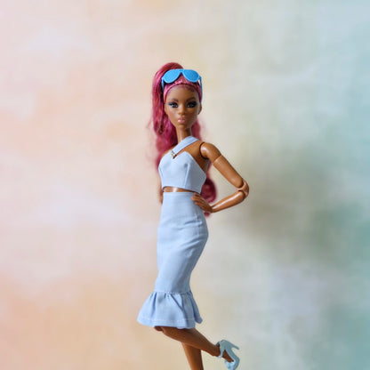 Dark-skinned Barbie doll with pink hair models pastel blue asymmetrical crop top and ruffle hem skirt.
