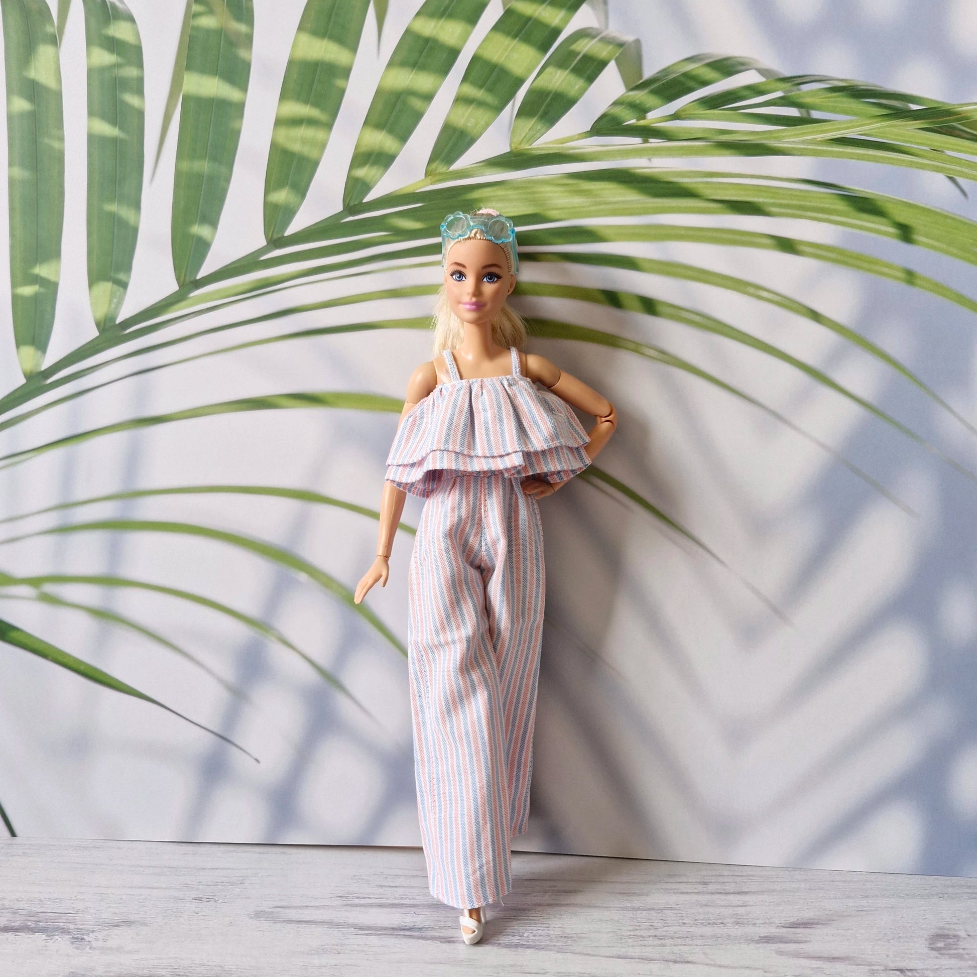 Barbie doll in pastel striped jumpsuit with ruffle top, standing by large palm leaf.