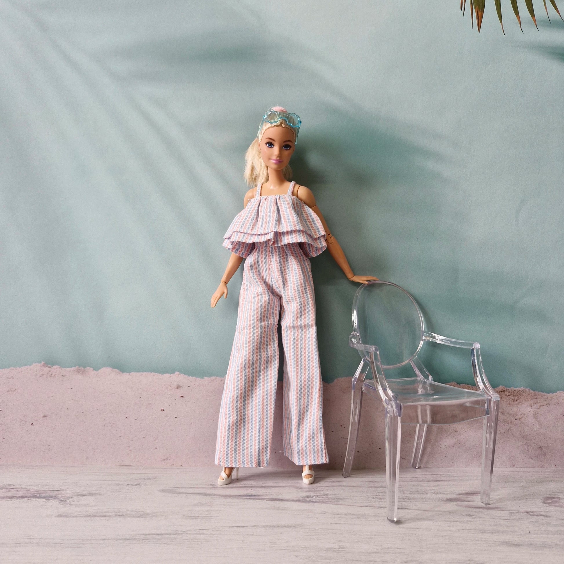Barbie doll in striped jumpsuit standing next to a clear miniature chair, beach theme.