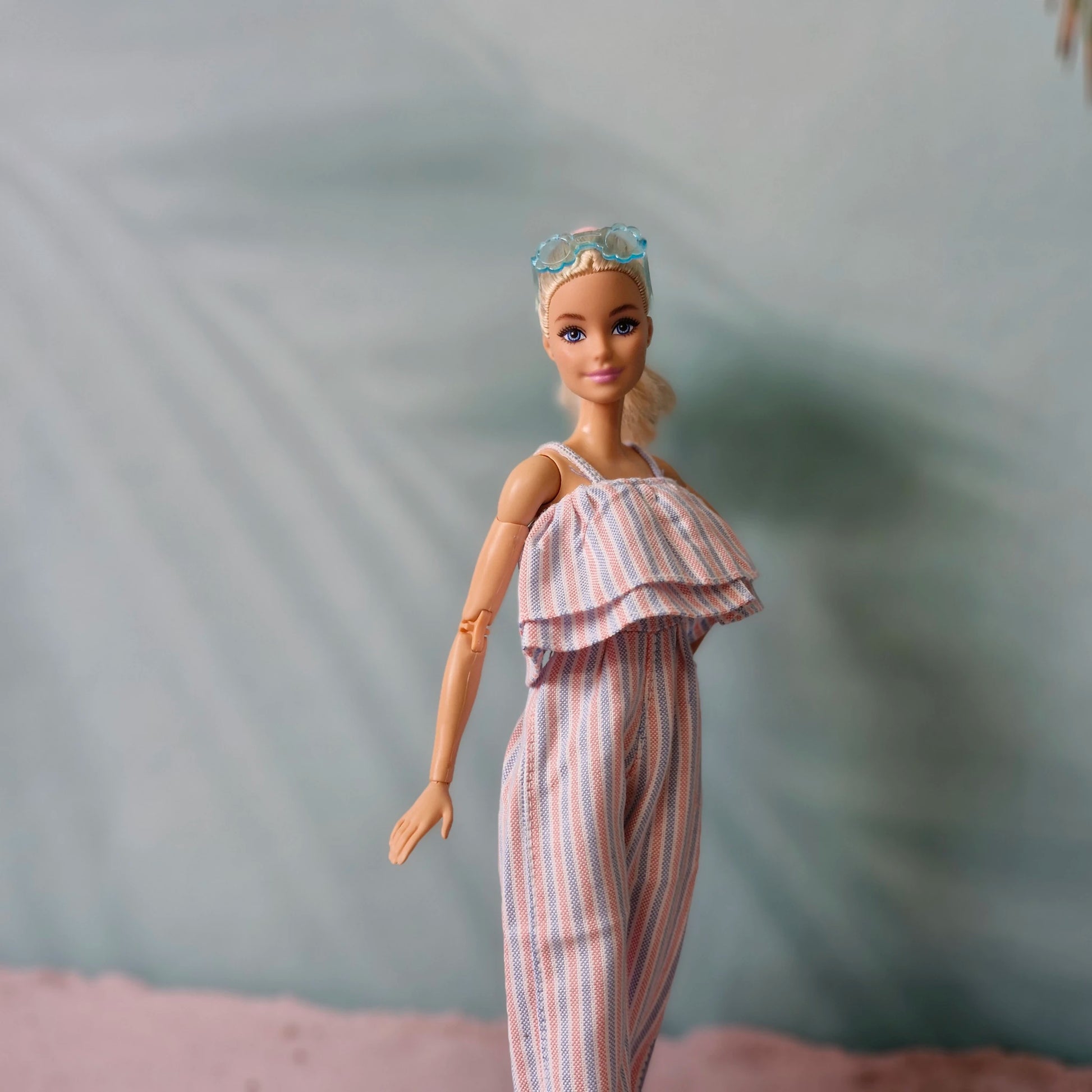 Blonde Barbie doll wearing striped ruffle jumpsuit and blue sunglasses on head.