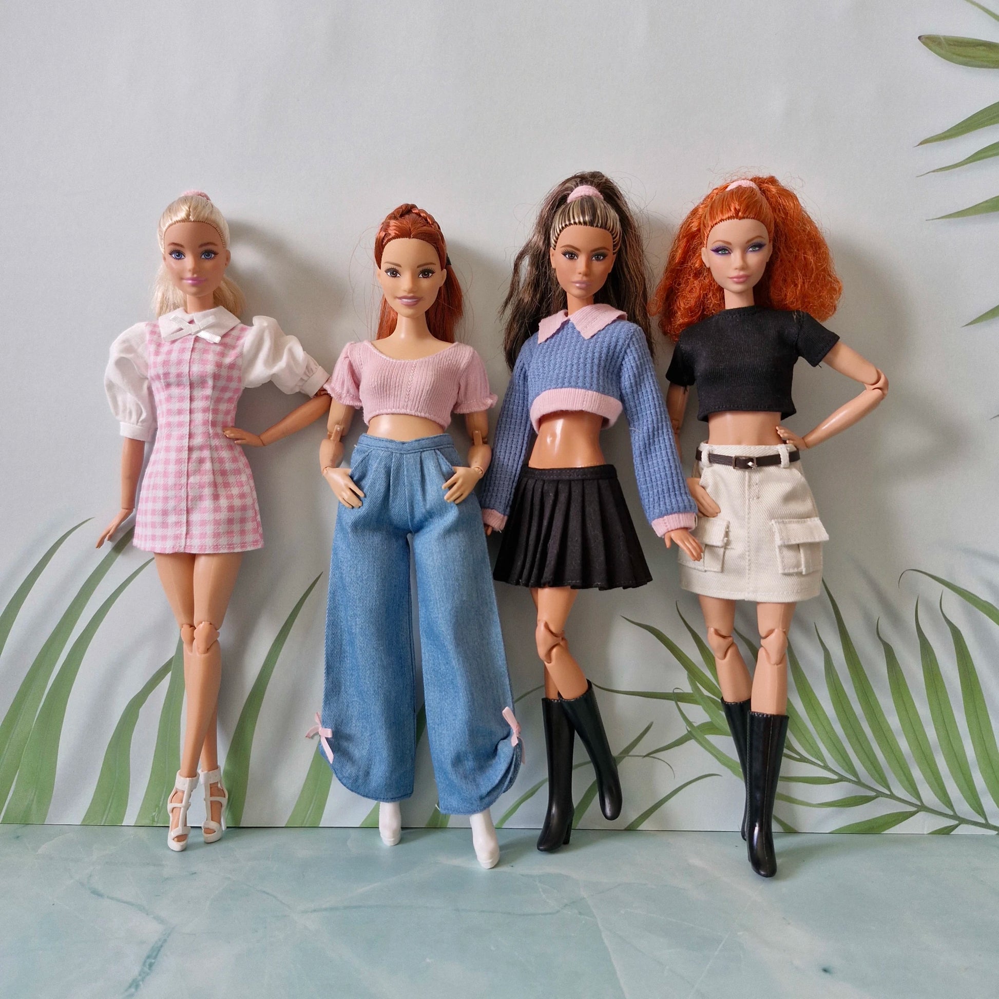 Group photo of four stylishly dressed Barbie dolls in modern fashion clothes against a light blue and green leaf background.