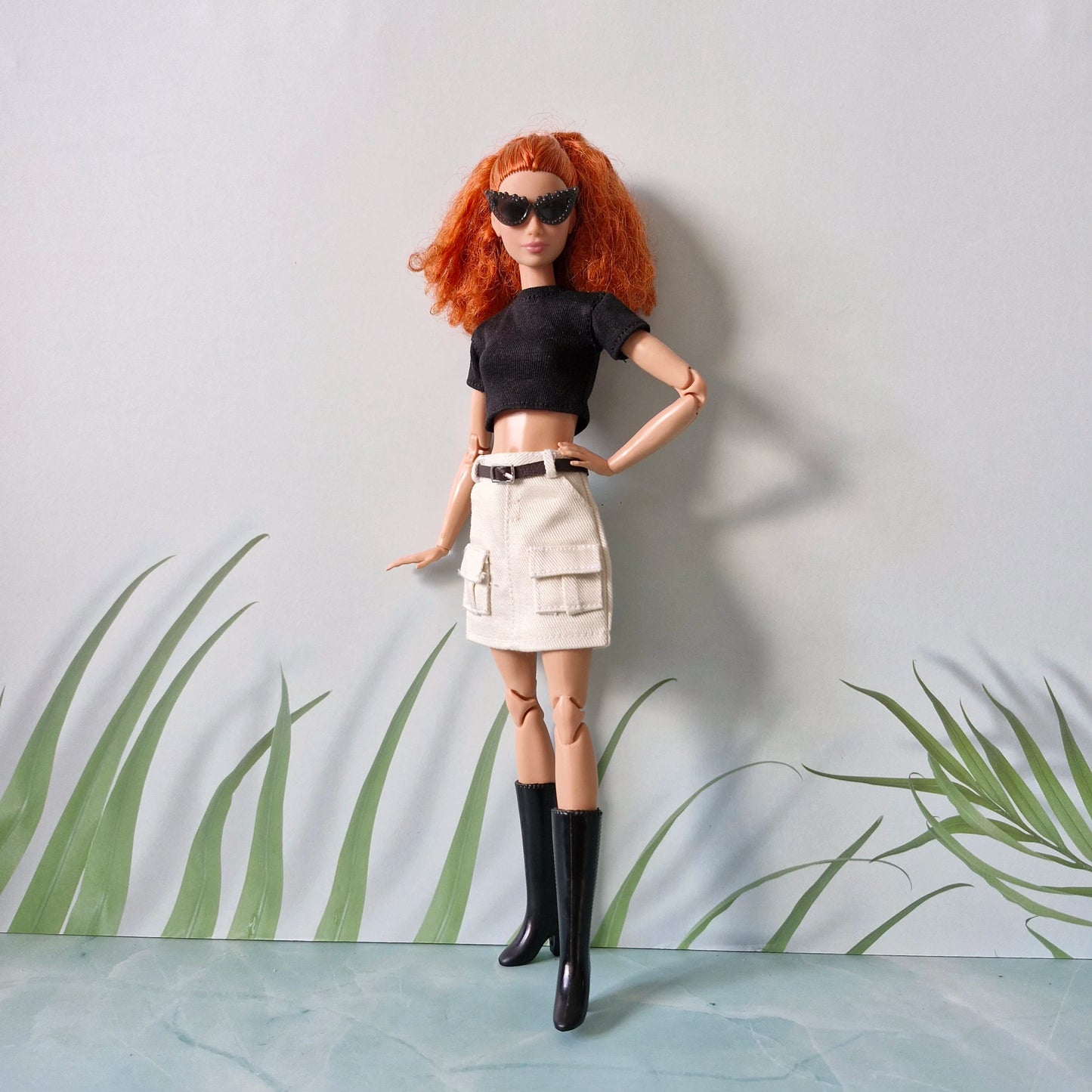 Red-haired Barbie doll wearing a black crop top, white cargo mini skirt, black boots, and sunglasses, posing with hands on hips