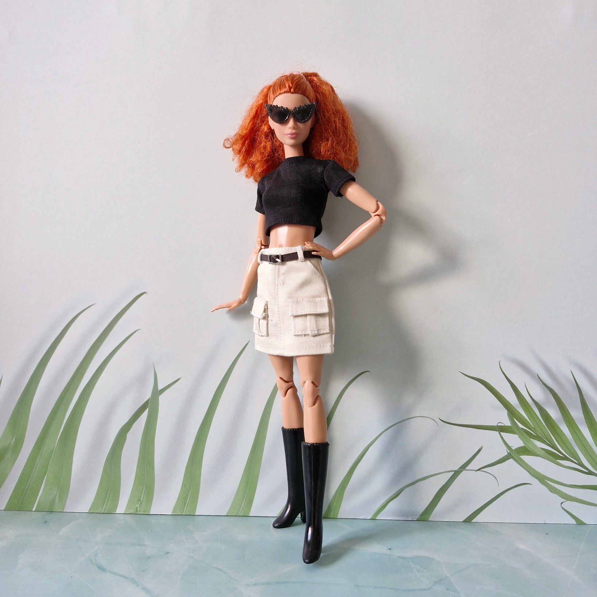 Red-haired Barbie doll wearing a black crop top, white cargo mini skirt, black boots, and sunglasses, posing with hands on hips