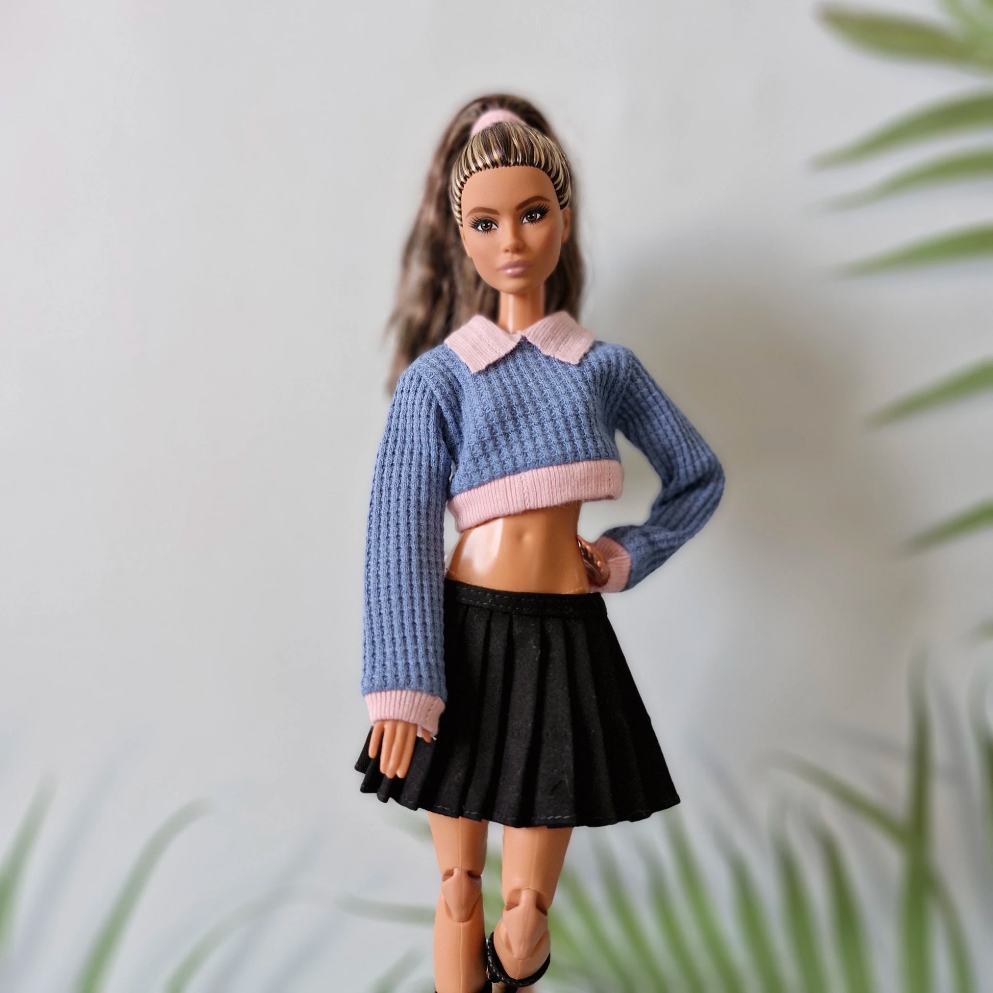 Brunette Barbie doll in a preppy blue crop sweater, black pleated skirt, and black boots, posing against a leafy background.