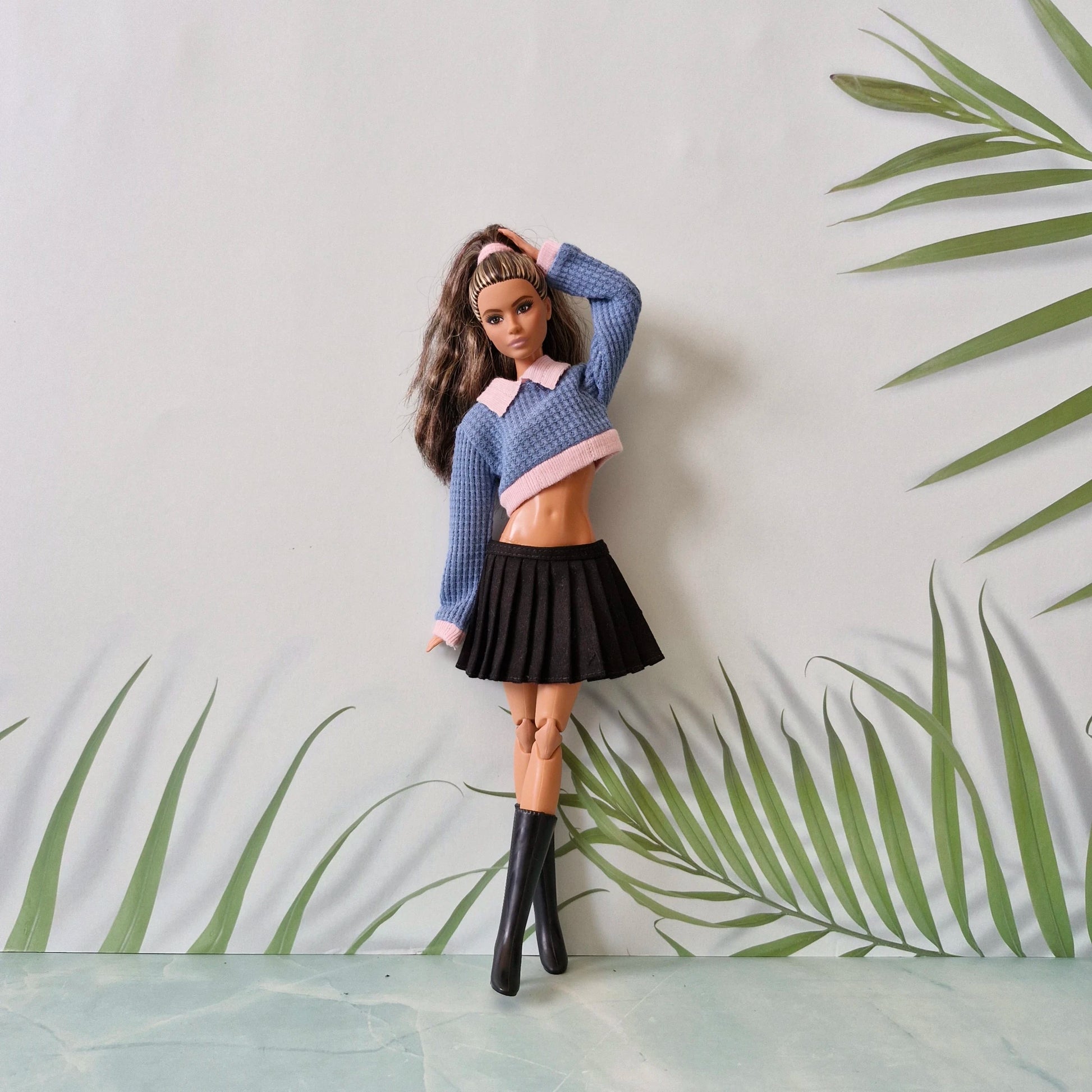 Close-up of a brunette Barbie doll in a blue and pink collared crop sweater and black pleated skirt.
