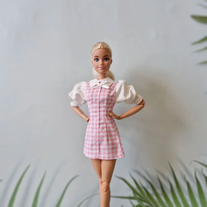 Blonde Barbie doll in a pink and white gingham pinafore dress over a white puff-sleeve blouse, posing with hands on hips.
