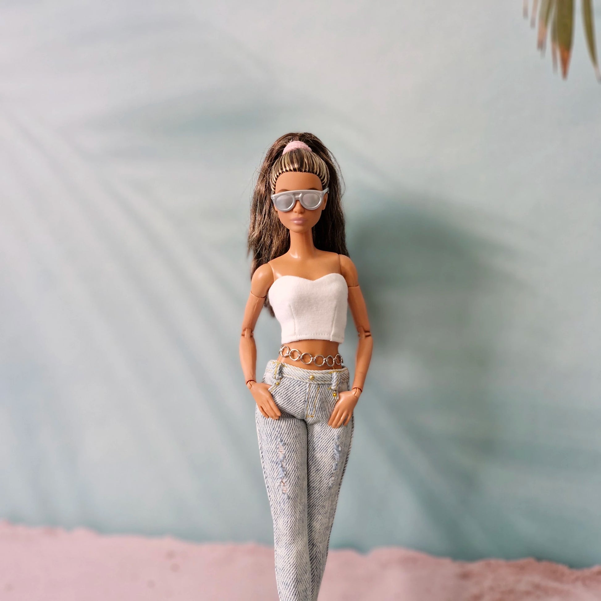 Brunette Barbie doll wearing sunglasses, a white tube top, and distressed jeans with a chain belt detail.