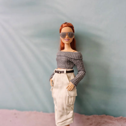 Barbie doll wearing sunglasses, a gray off-shoulder top, and beige pants against a plain background