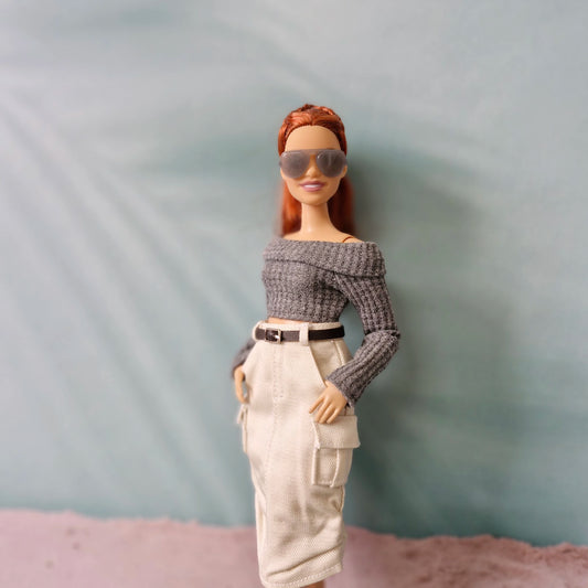 Barbie doll wearing sunglasses, a gray off-shoulder top, and beige pants against a plain background