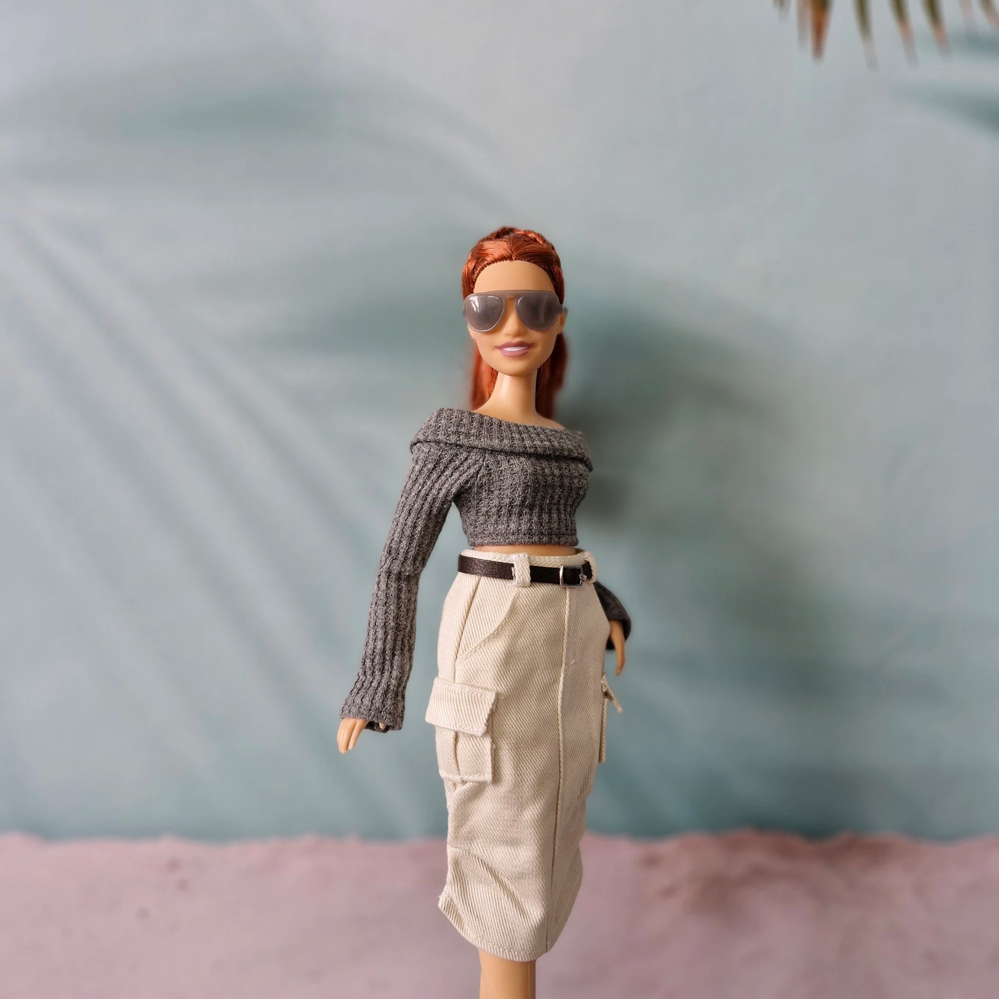 Barbie doll wearing a gray sweater and beige pants with sunglasses against a blurred background