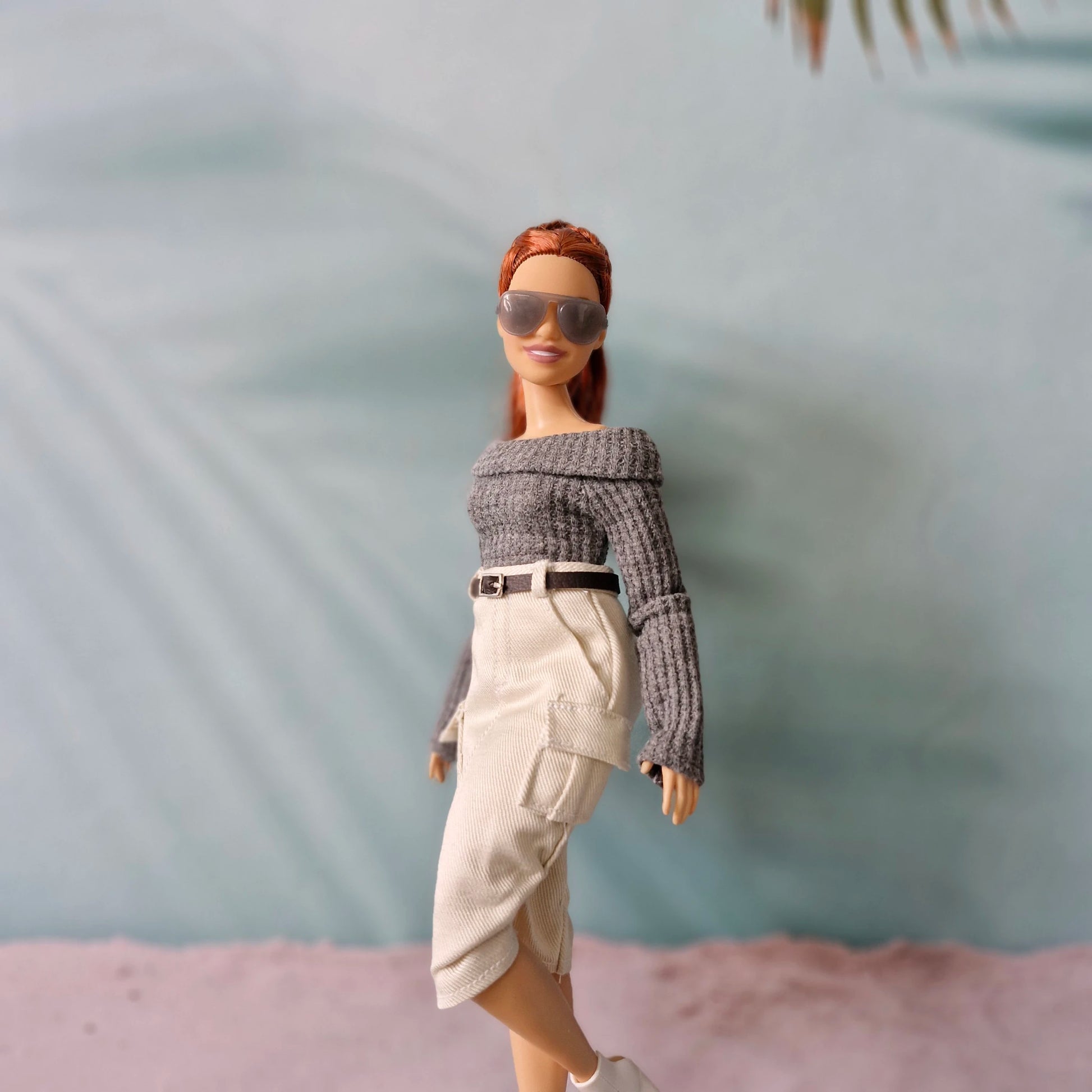 Barbie doll wearing sunglasses and a gray sweater with a beige skirt against a blurred background