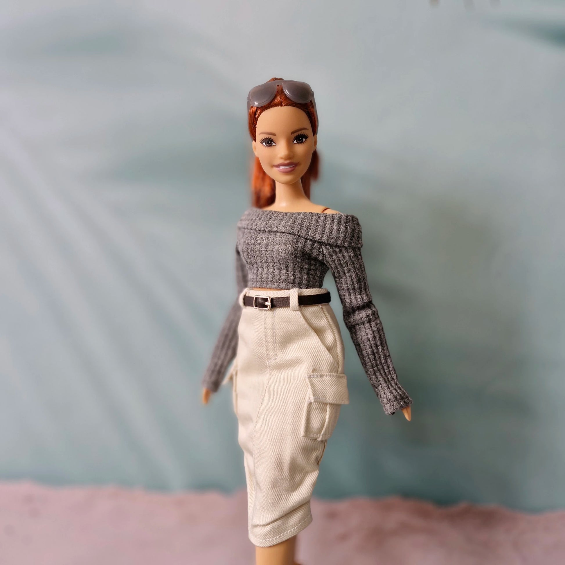 Barbie doll wearing a gray off-shoulder top and beige pants against a plain background