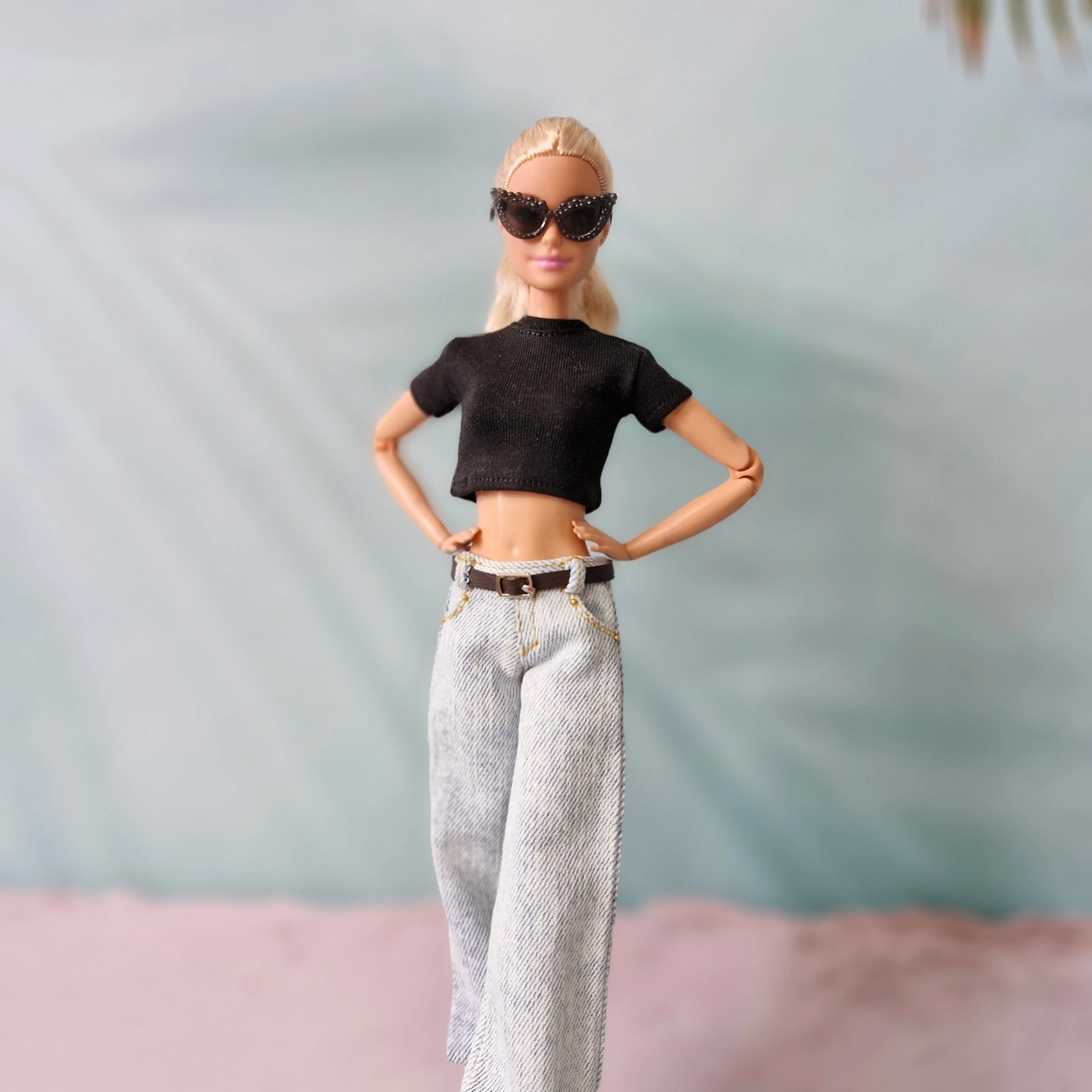 Barbie doll wearing a black crop top and light-colored pants with sunglasses against a neutral background