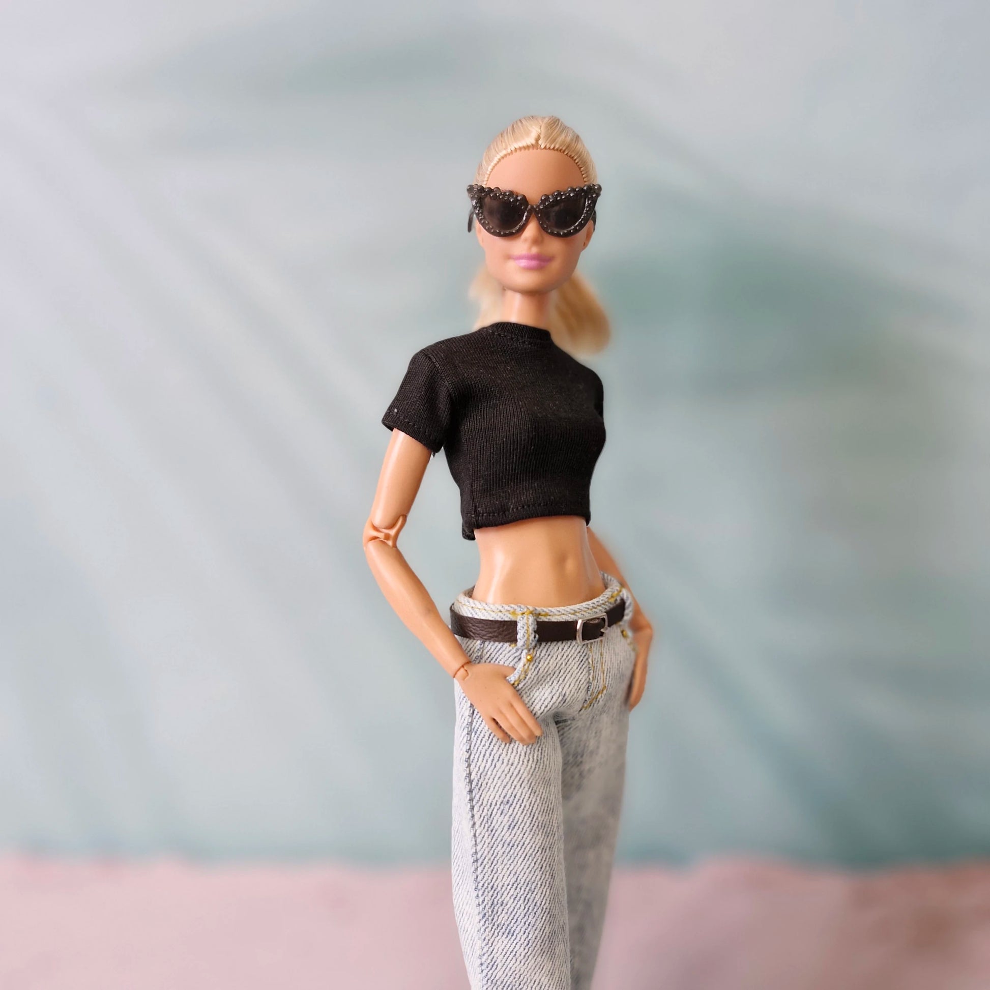 Barbie doll wearing sunglasses, black top, and gray pants against a plain background