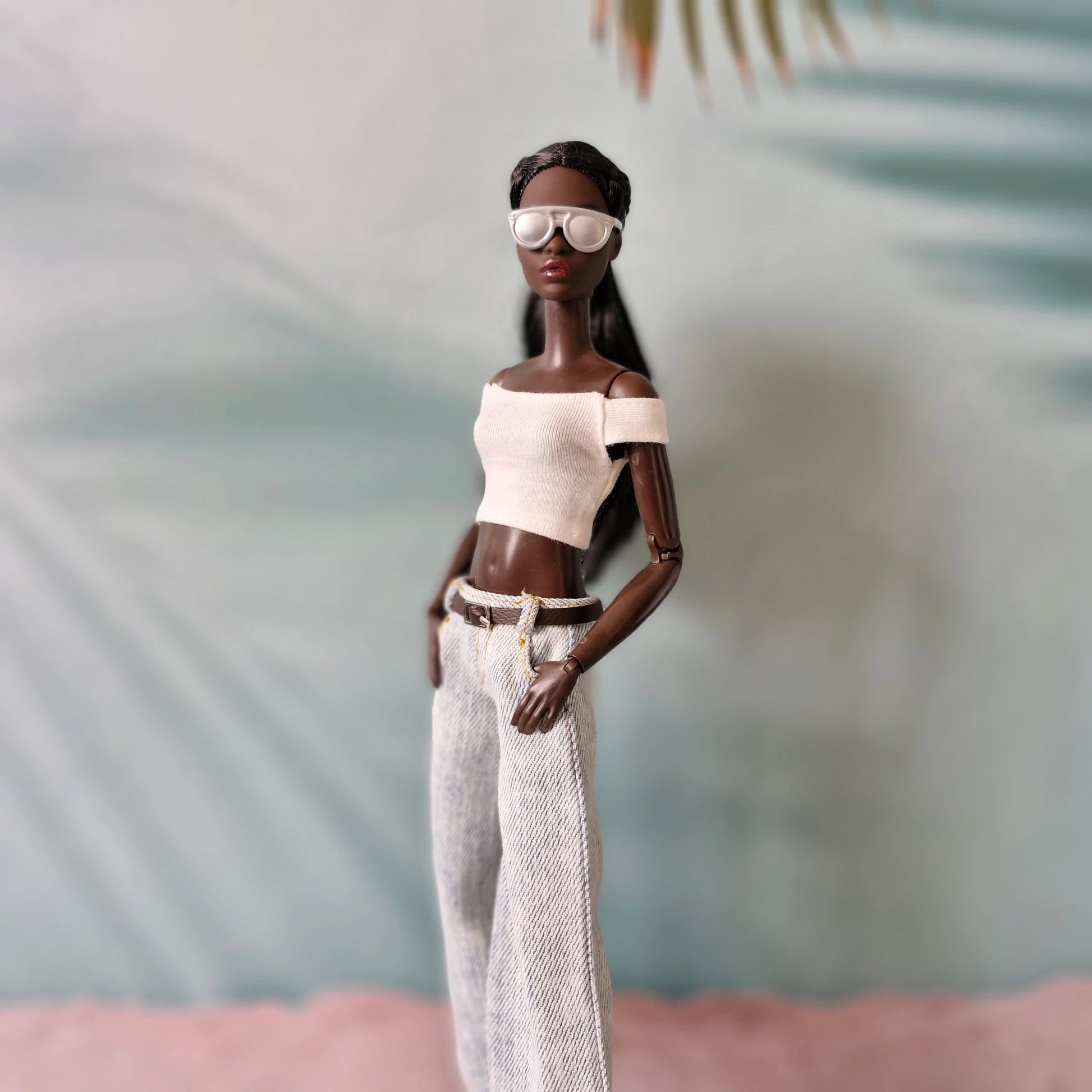 Doll wearing a white top and pants with sunglasses against a blurred natural background