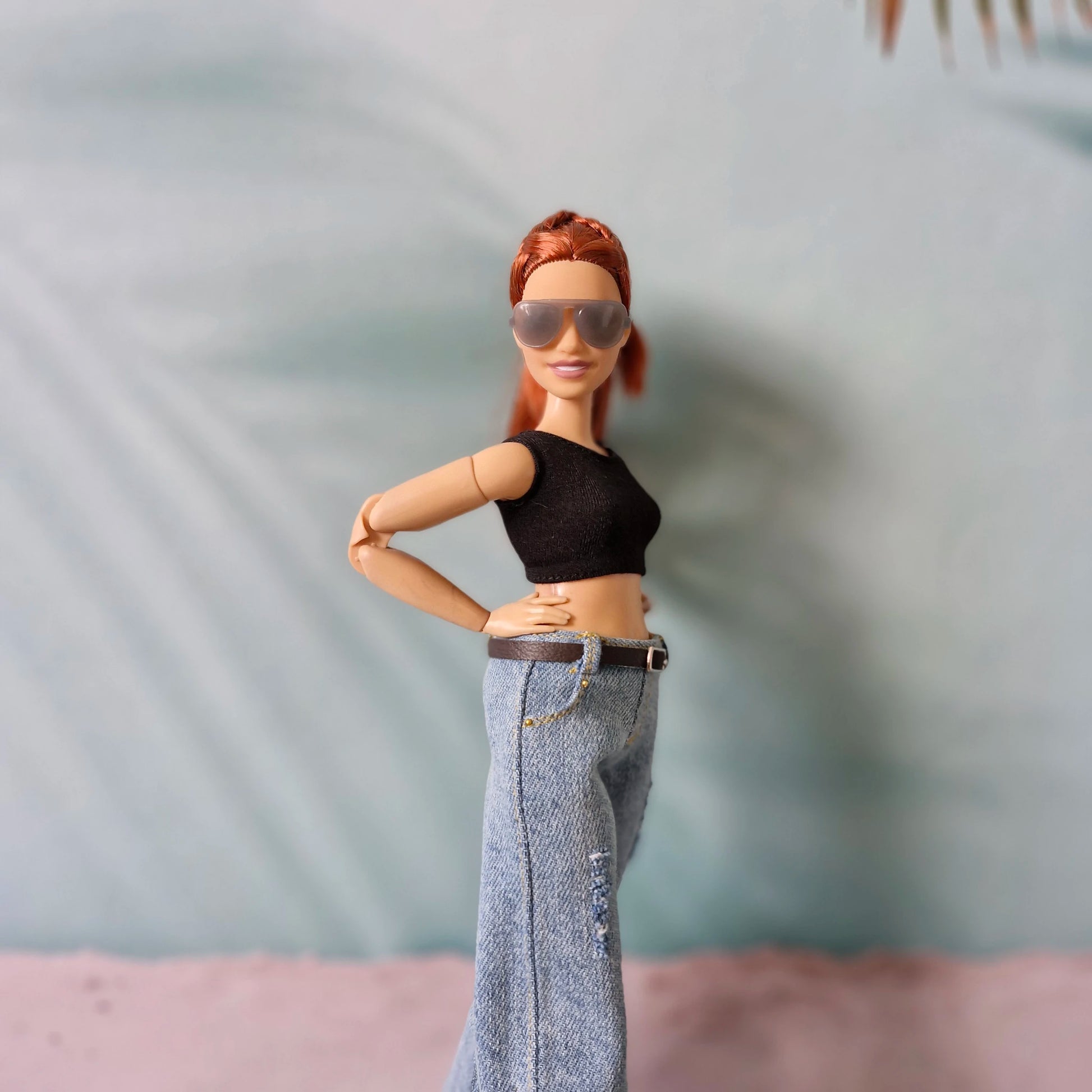 Barbie doll with red hair, sunglasses, black top, and blue jeans against a blurred background