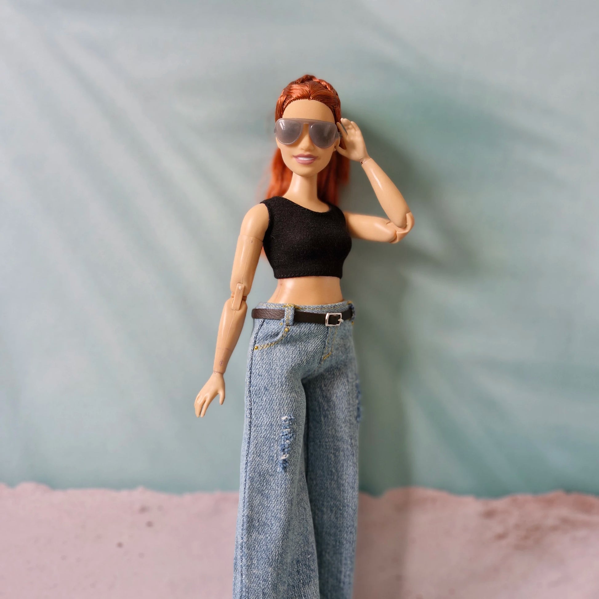 Doll with red hair wearing sunglasses and a black top with blue jeans against a neutral background