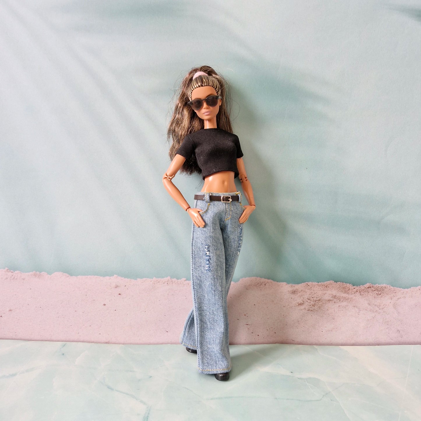 Barbie doll wearing a black crop top and gray flared pants with sunglasses on a light blue background.