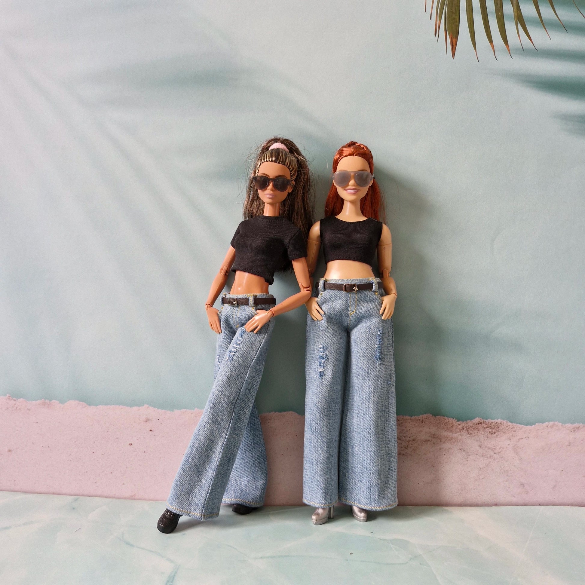 Two dolls wearing black tops and wide-leg jeans against a light blue wall.