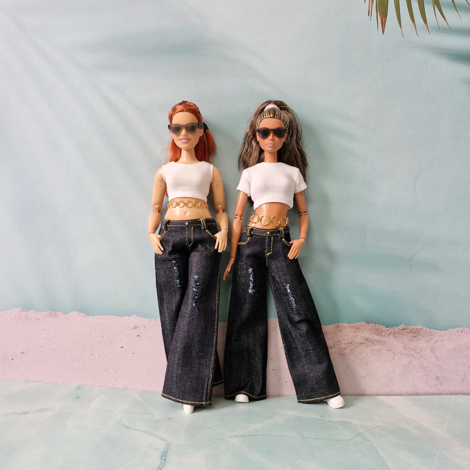 Two dolls wearing matching outfits with white tops and dark jeans against a light blue background.