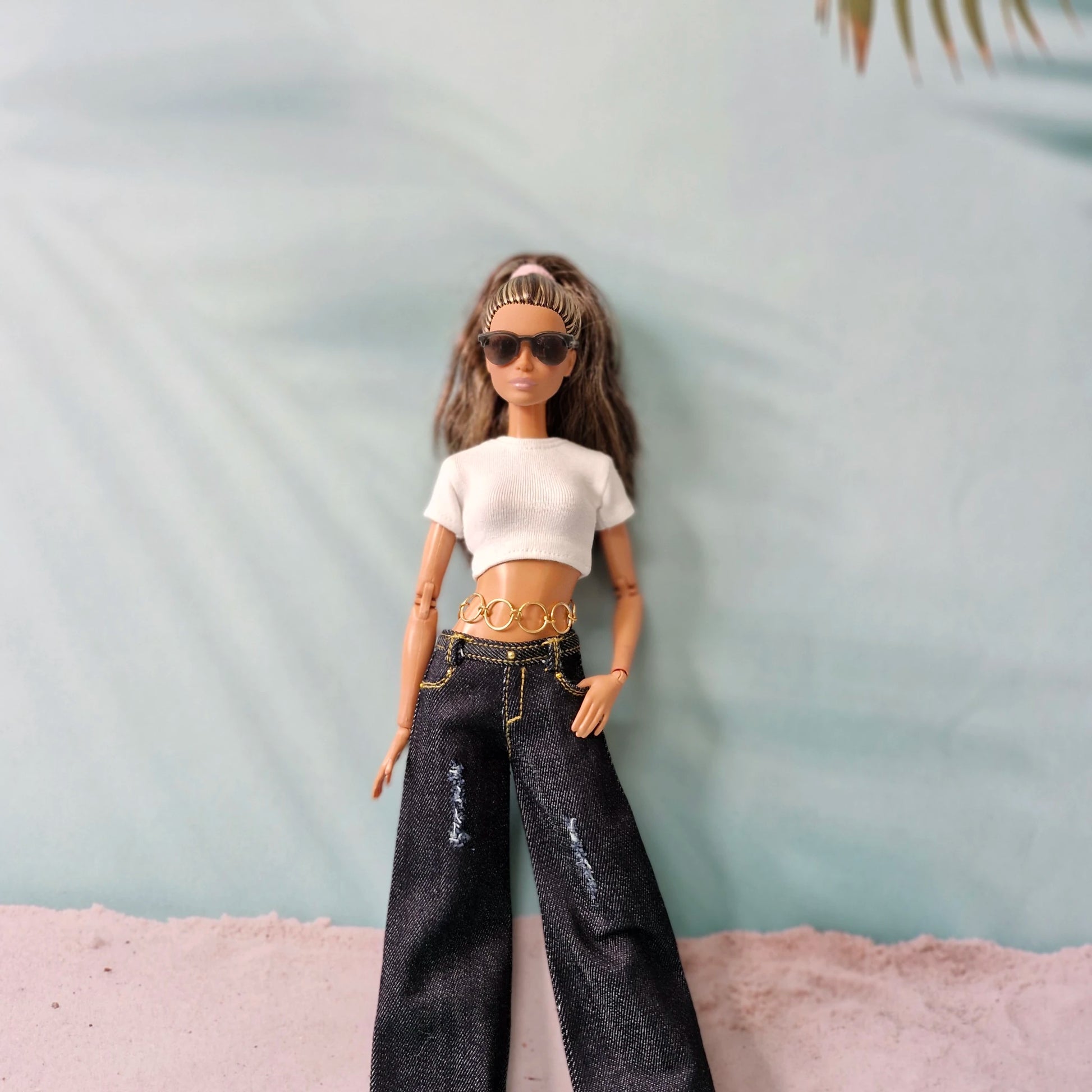 Barbie doll wearing a white top, black jeans, and sunglasses against a light blue background.