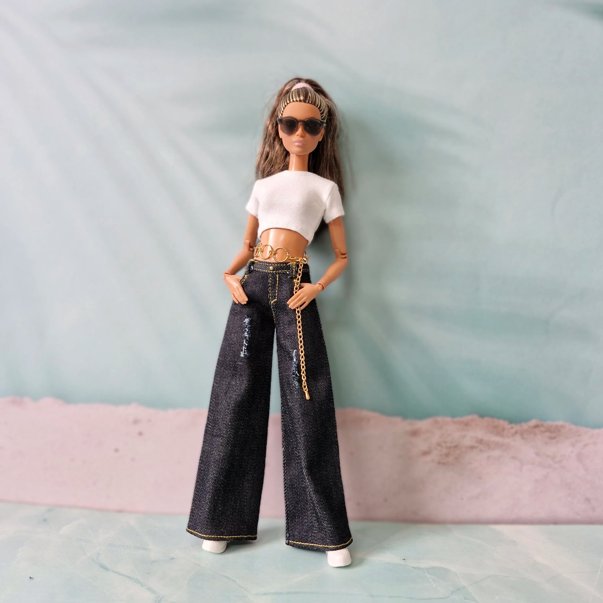 Barbie doll wearing a white top and black jeans with a gold chain, standing against a light blue background.