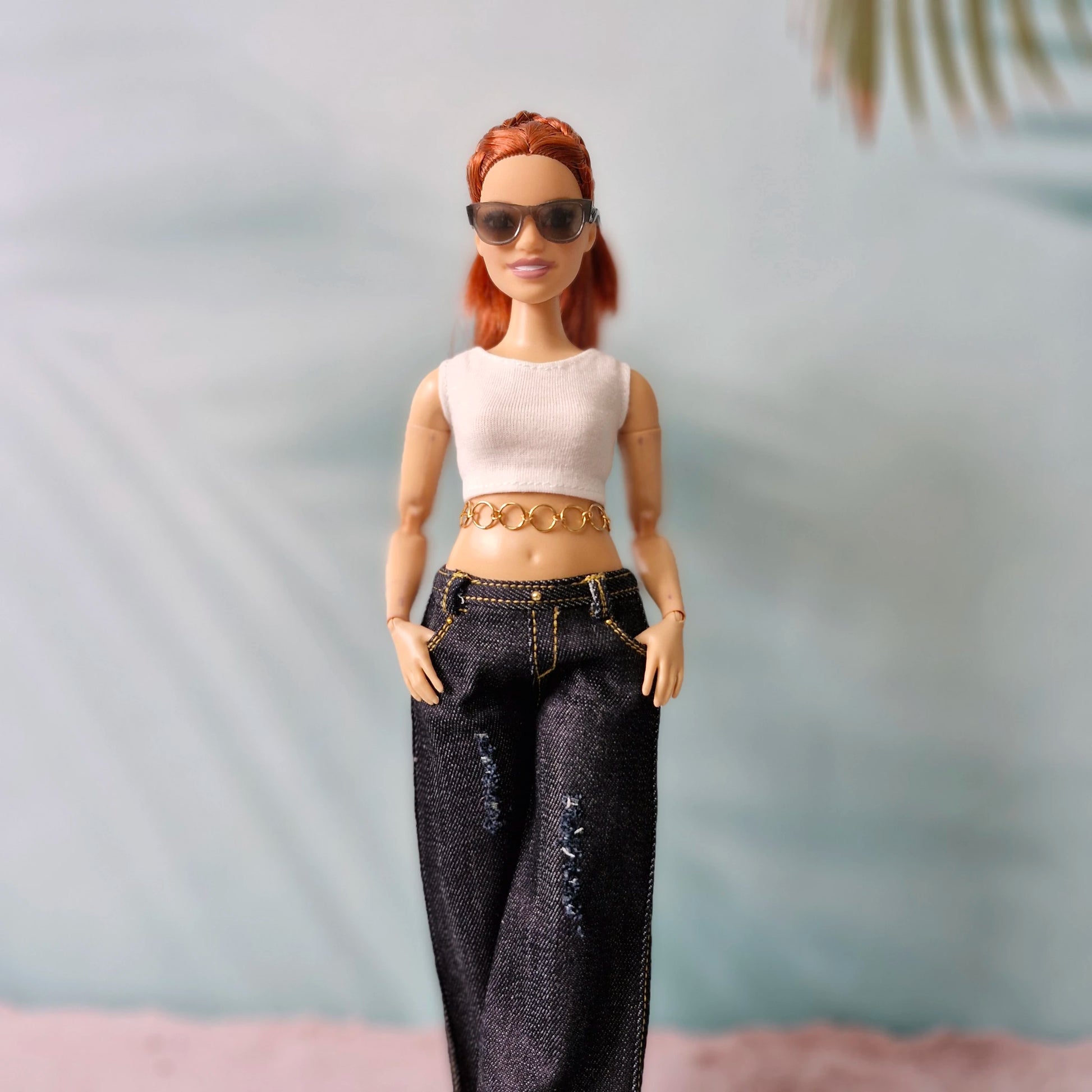 Doll with red hair wearing sunglasses, a white top, and black jeans against a neutral background