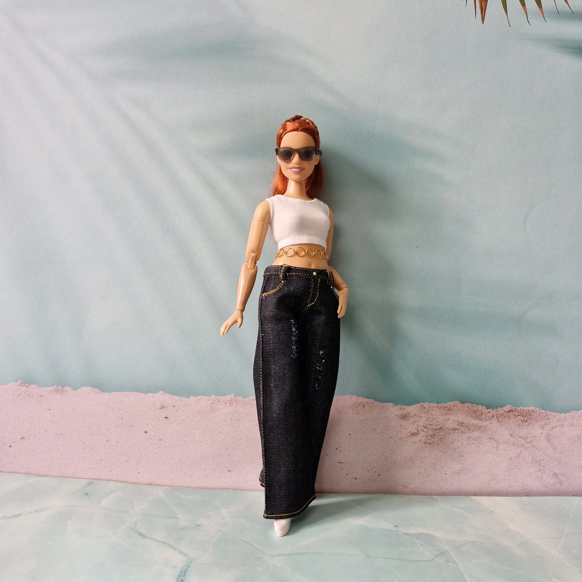 Barbie doll with red hair, sunglasses, white top, and black pants standing on a textured surface.
