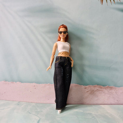 Barbie doll with red hair, sunglasses, white top, and black pants standing on a textured surface.