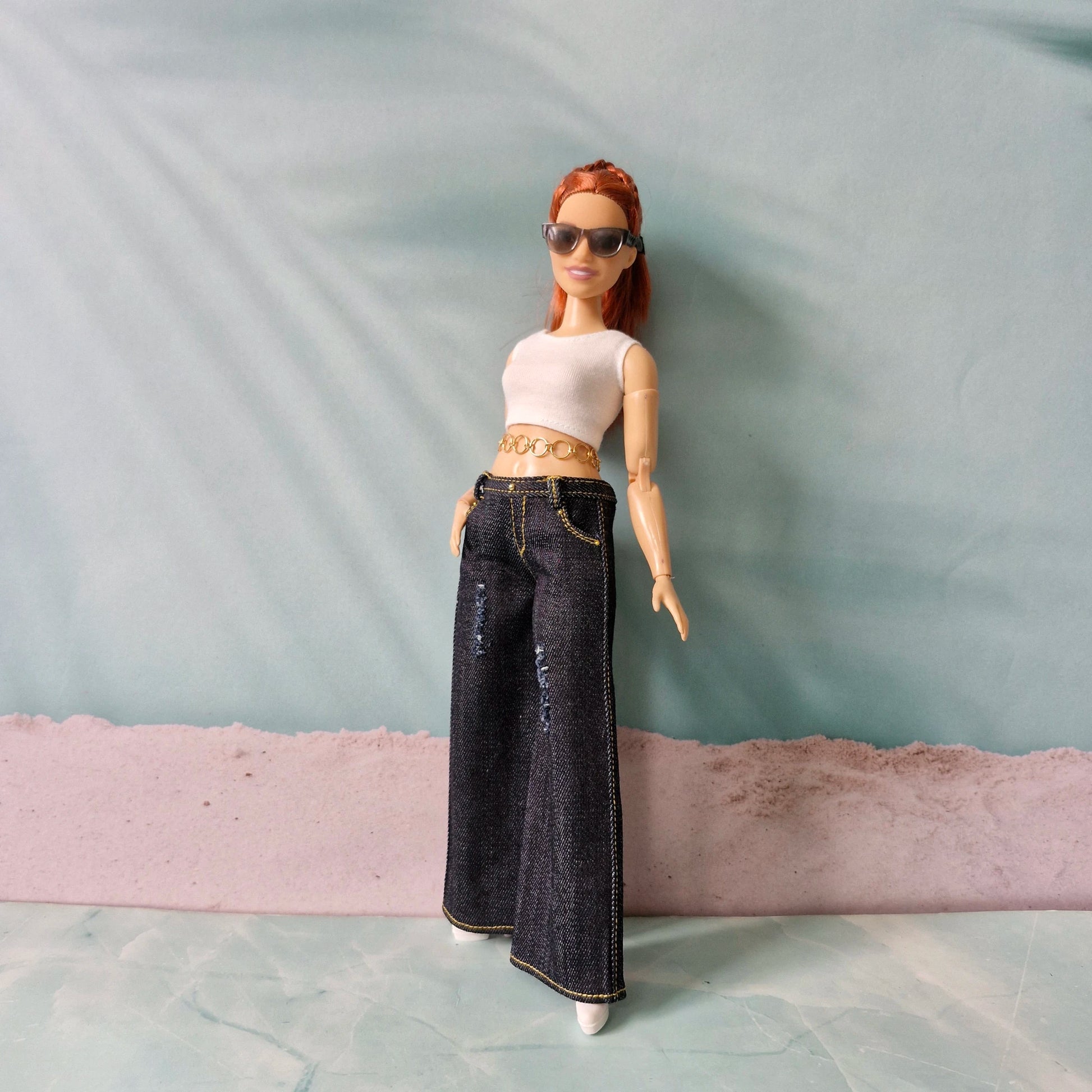 Doll wearing a white top and blue jeans with sunglasses on a plain background