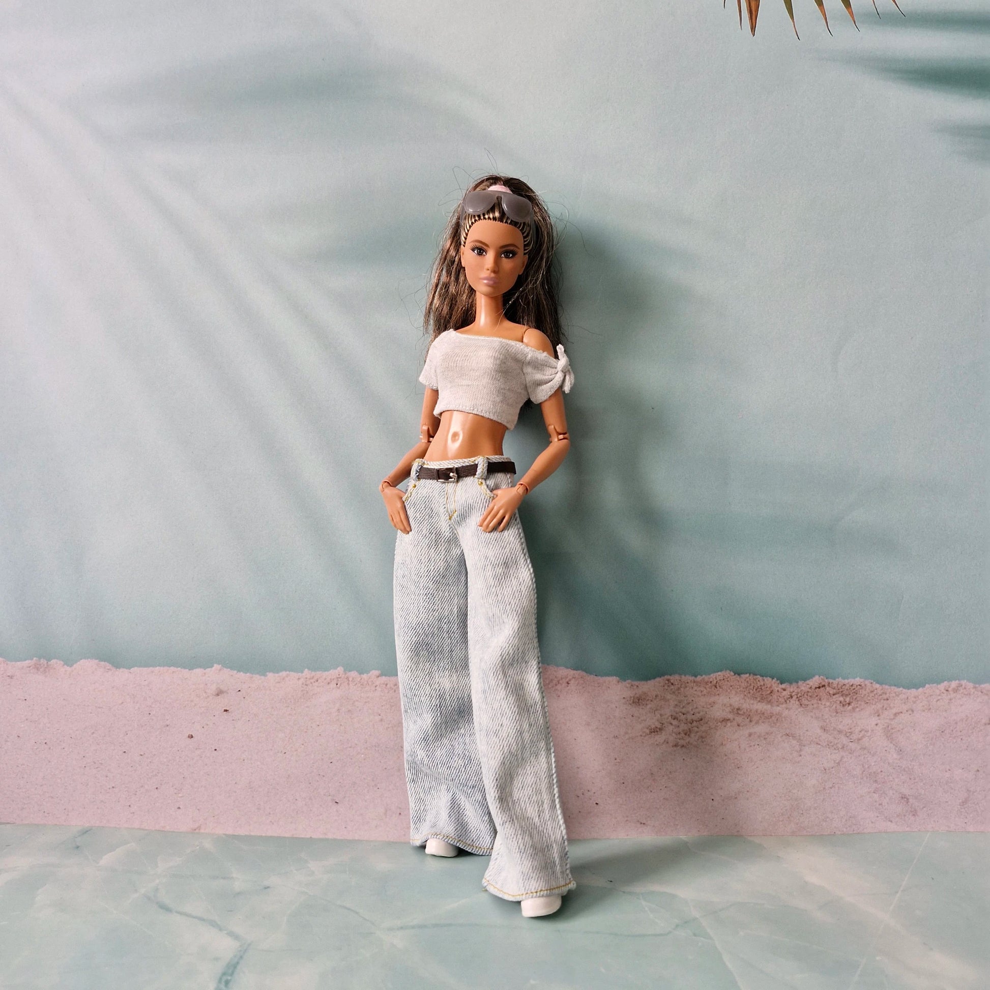 Barbie doll wearing a white off-shoulder top and light gray pants against a textured wall.