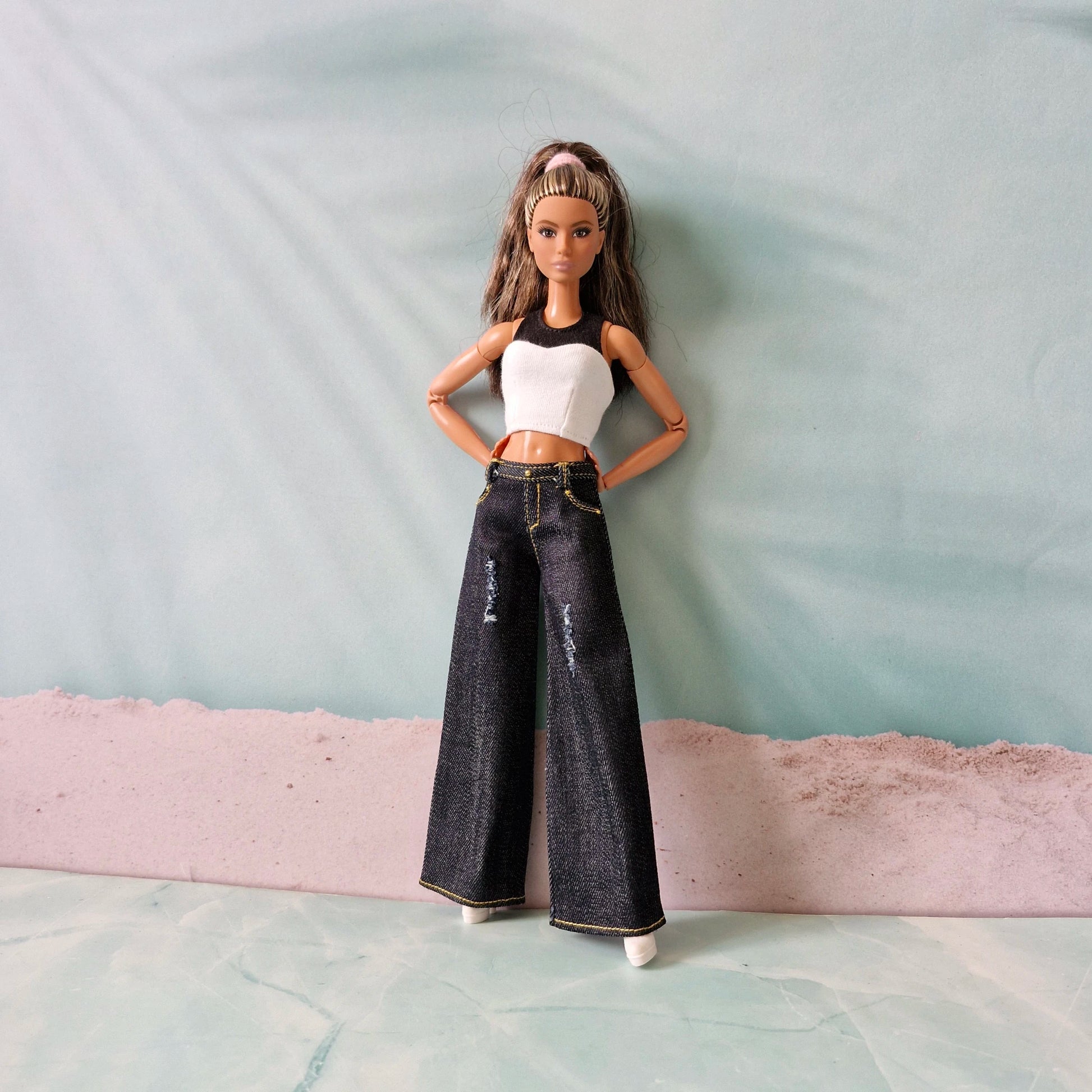 Barbie doll wearing a white top and black jeans standing on a textured surface with a light blue background.
