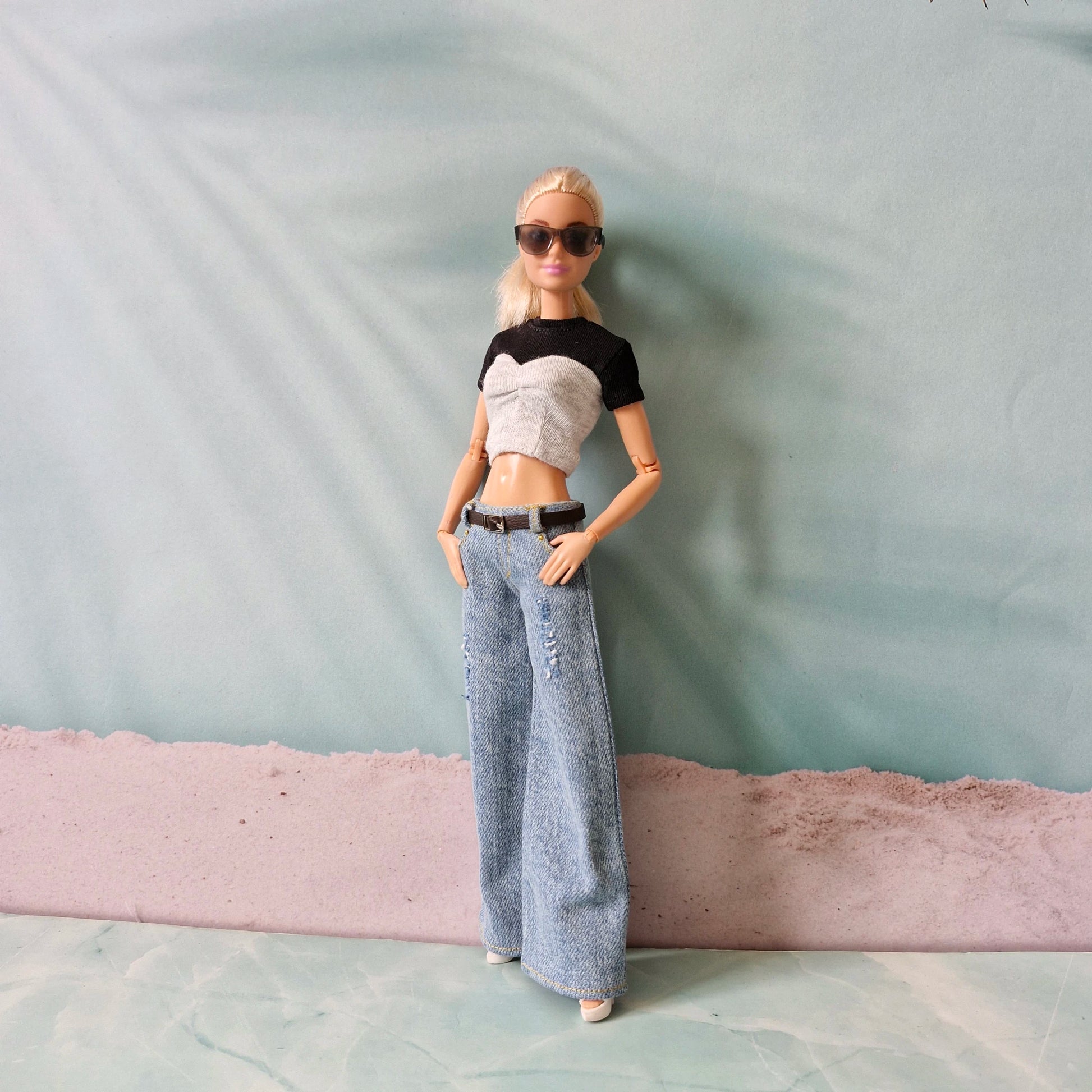 Barbie doll wearing a black and white crop top, blue jeans, and sunglasses against a light blue background.