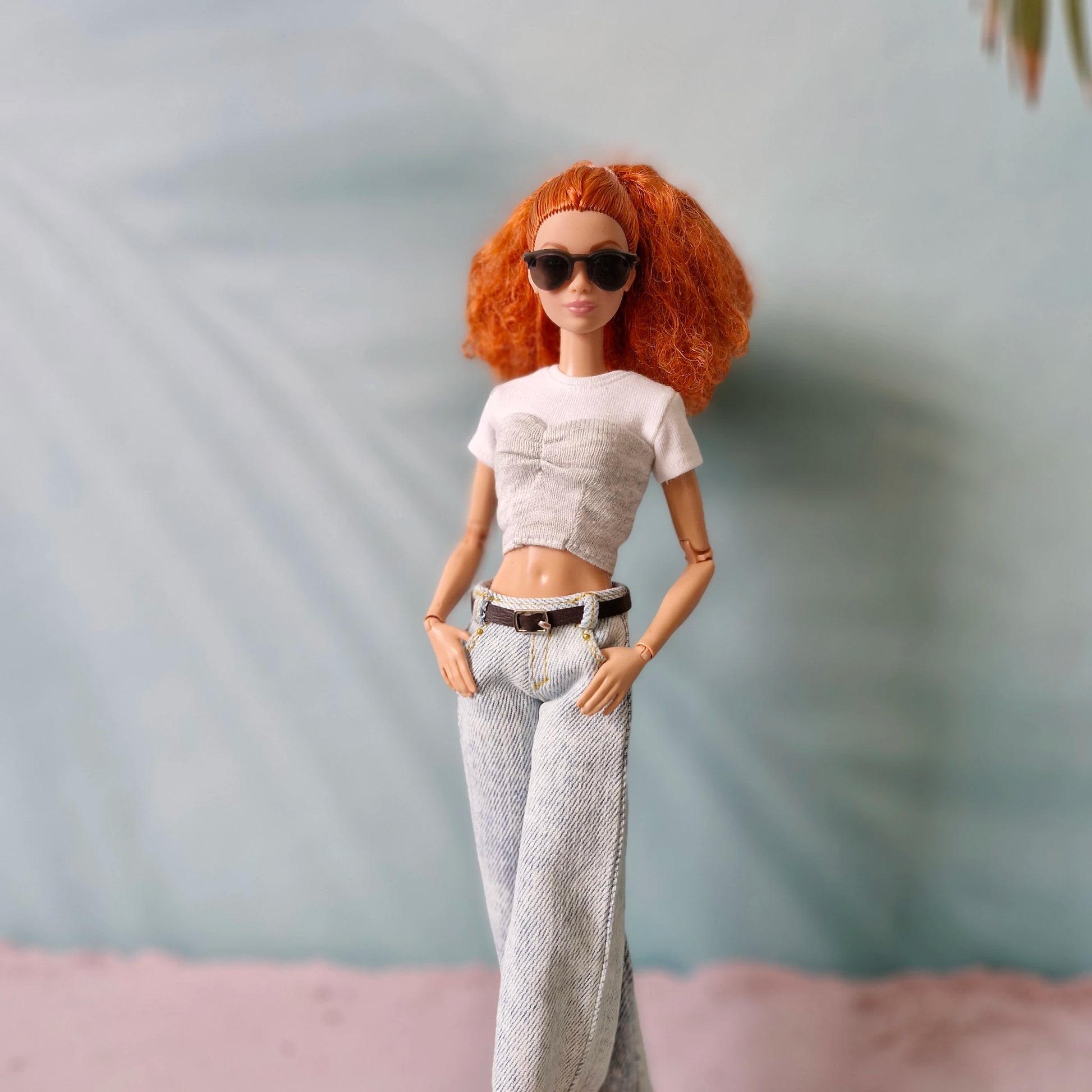 Doll with red hair wearing sunglasses, a white top, and gray pants against a neutral background