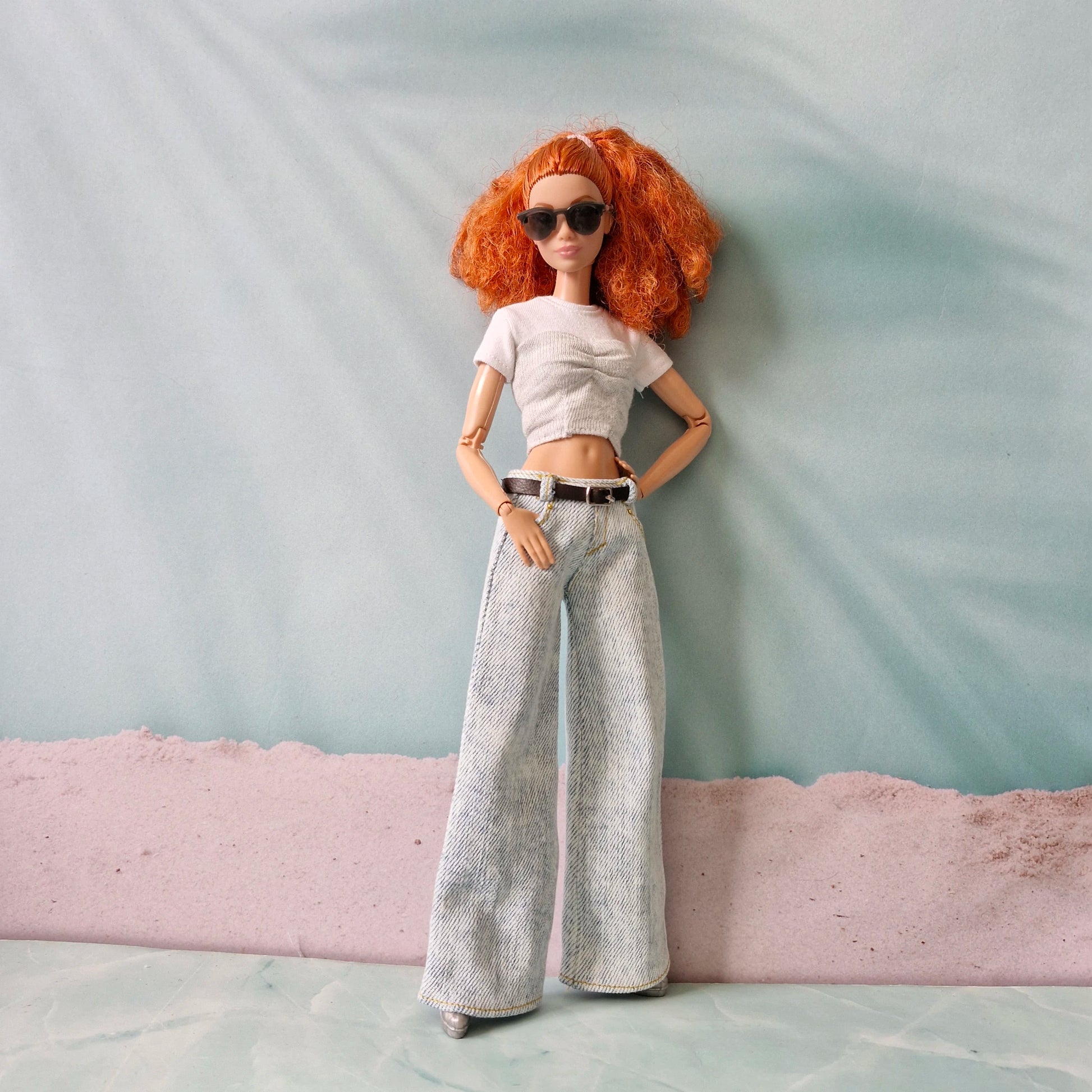 Doll with red hair wearing sunglasses, a white top, and gray pants against a light blue background.
